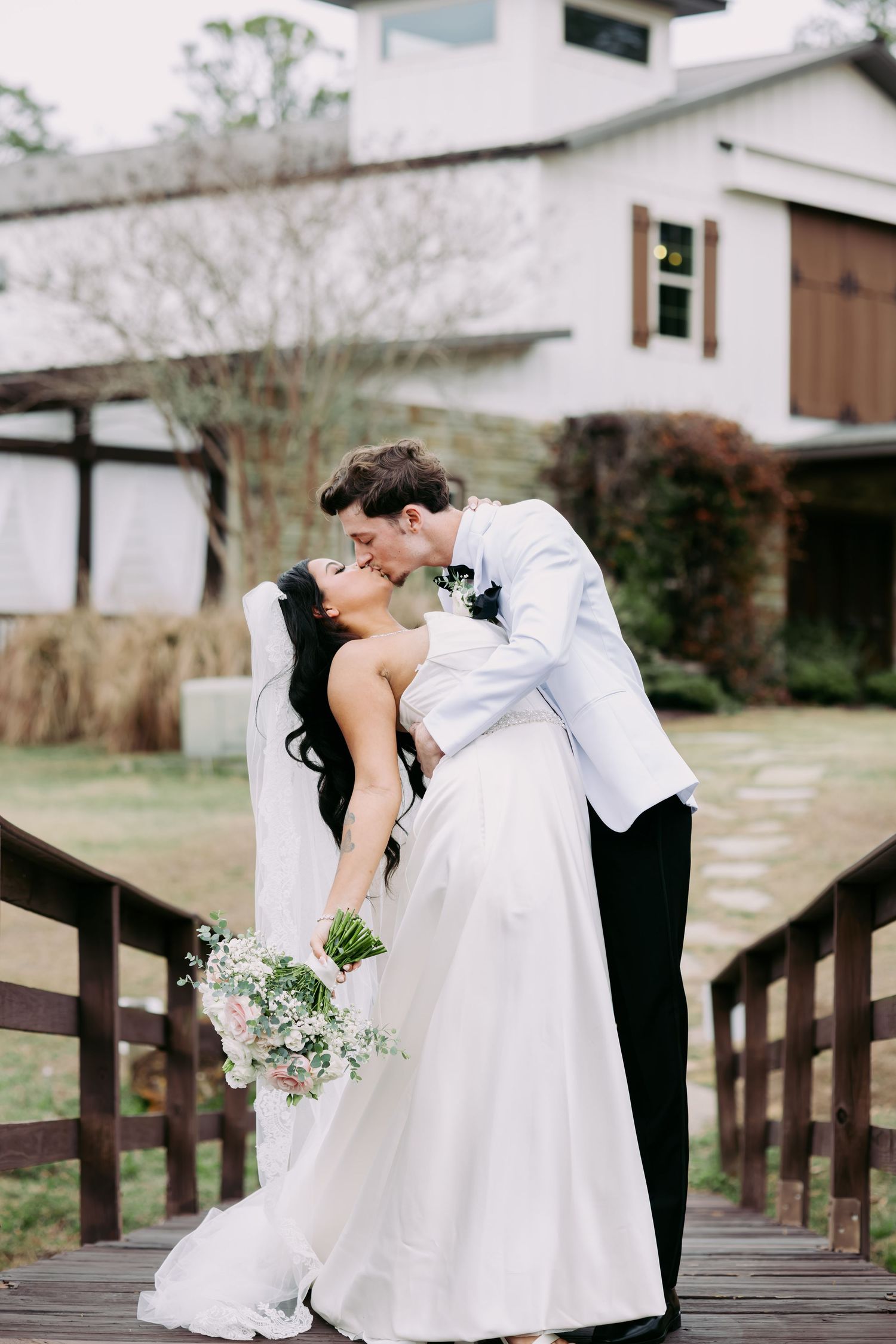 Groom in white tuxedo jacket dips and kisses bride on a wooden bridge at wedding venue.