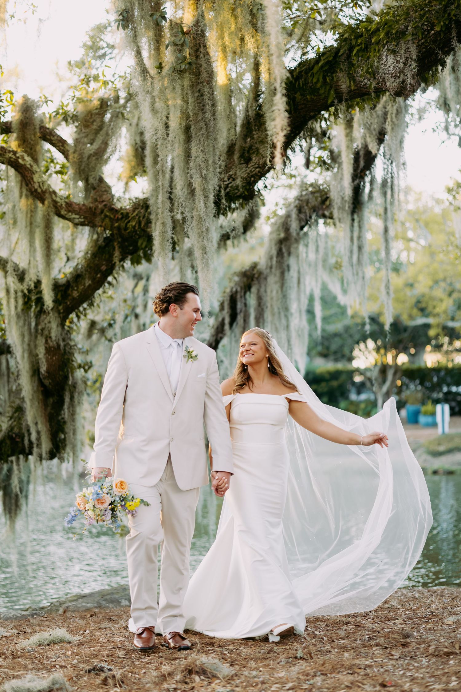 Joyful bride and groom walking hand-in-hand under Spanish moss-draped oak trees beside a pond.