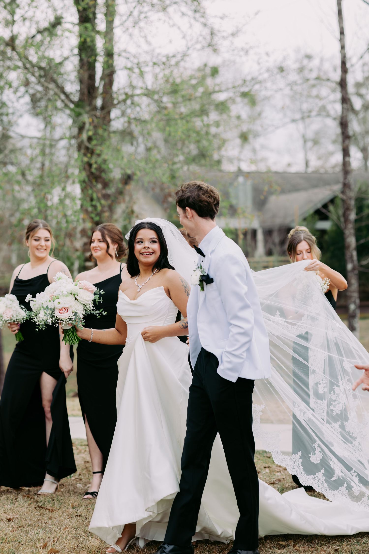 Groom lifting bride's lace-trimmed veil in the breeze while bridesmaids in black dresses look on outdoors.