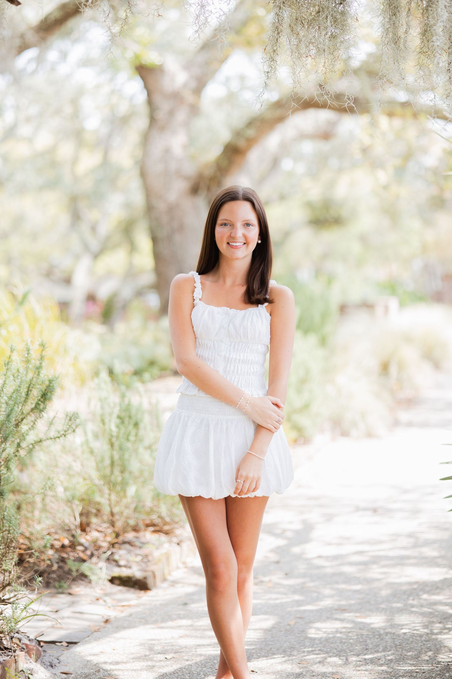 Smiling brunette woman in white sundress walking on a garden path under moss-draped oak trees.