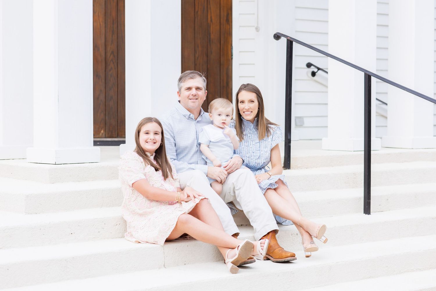 A family of four sits together on white steps in front of a wooden door, smiling in coordinating pastel outfits.