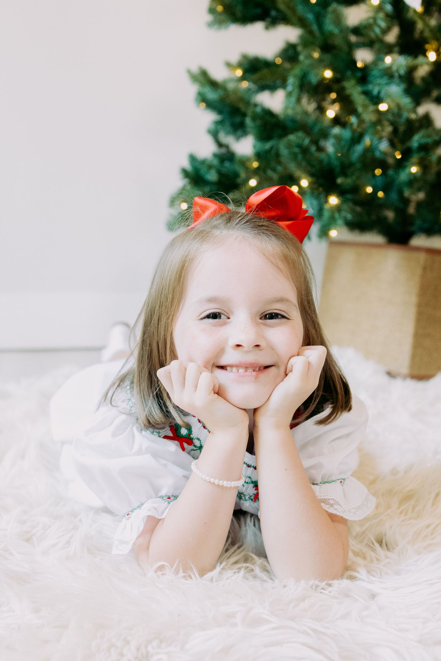 Smiling little girl with a red bow lying on a white fluffy rug in front of a glowing Christmas tree.