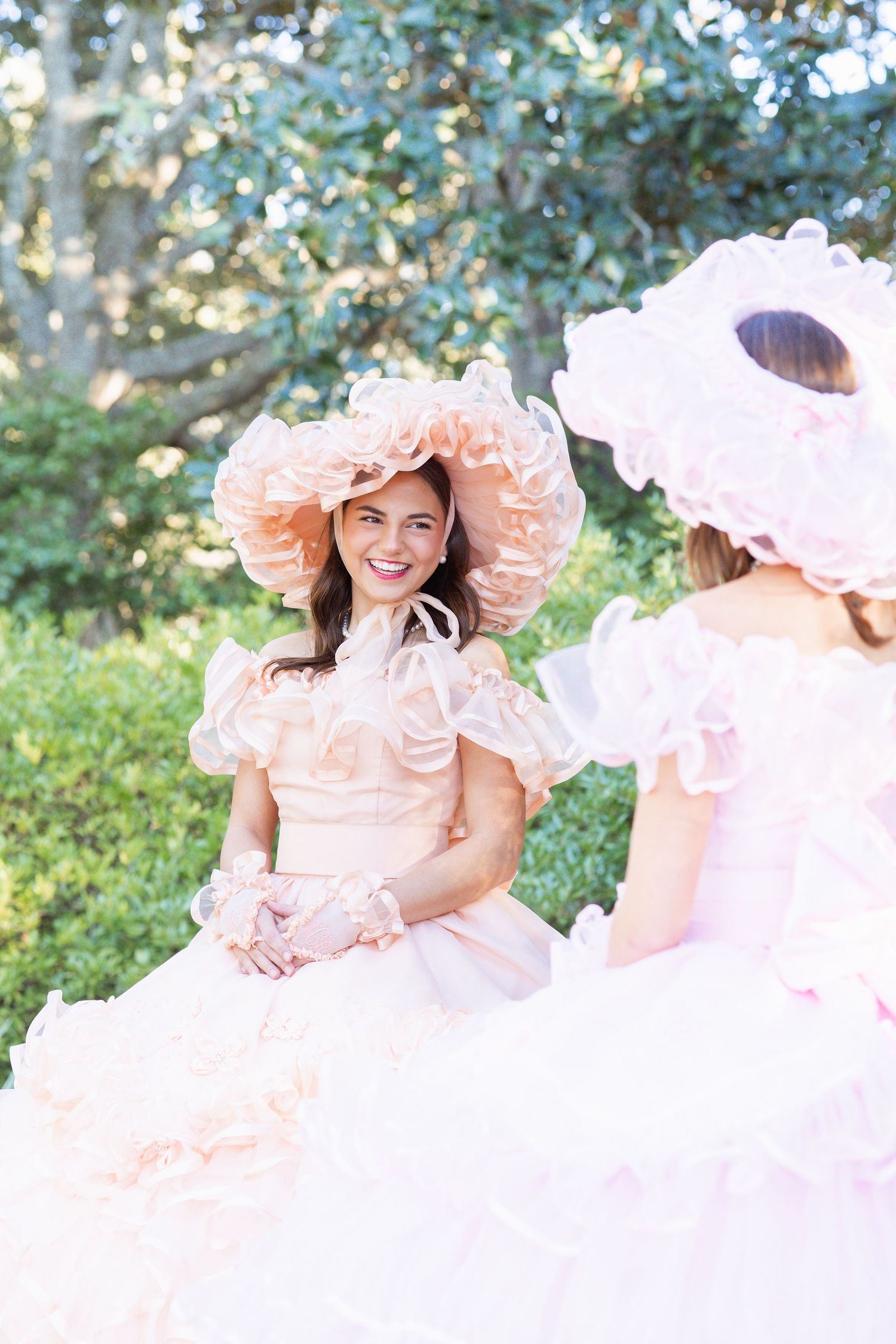 Two women in elegant pink ruffled ballgowns and floral hats conversing outdoors in a lush garden.