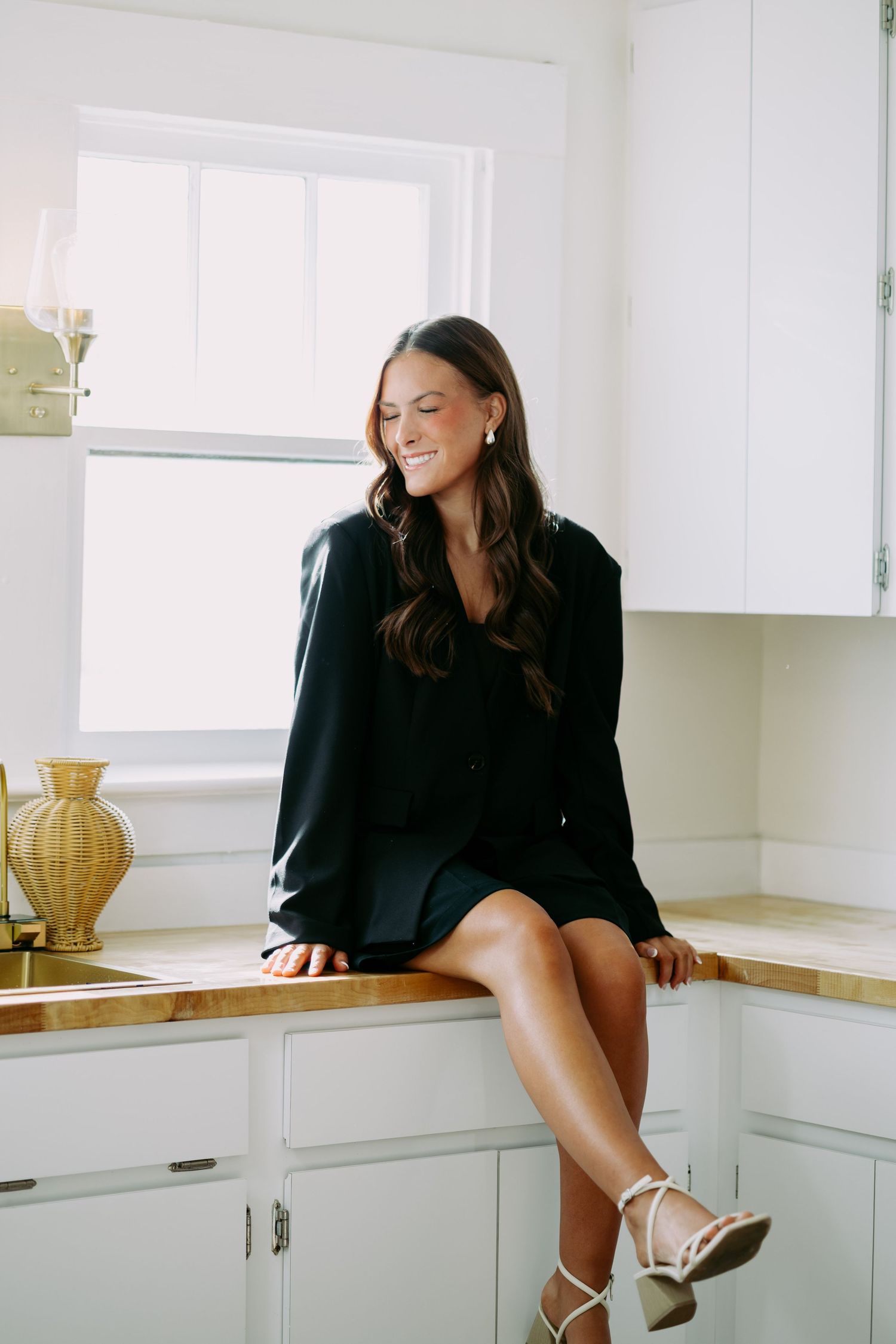 Smiling woman in black blazer dress sitting on kitchen counter near window in bright white kitchen.