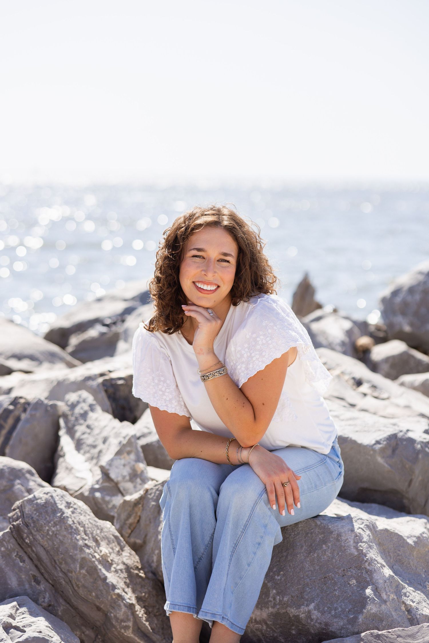 Smiling young woman with curly hair sitting on rocks by sparkling ocean on sunny day.