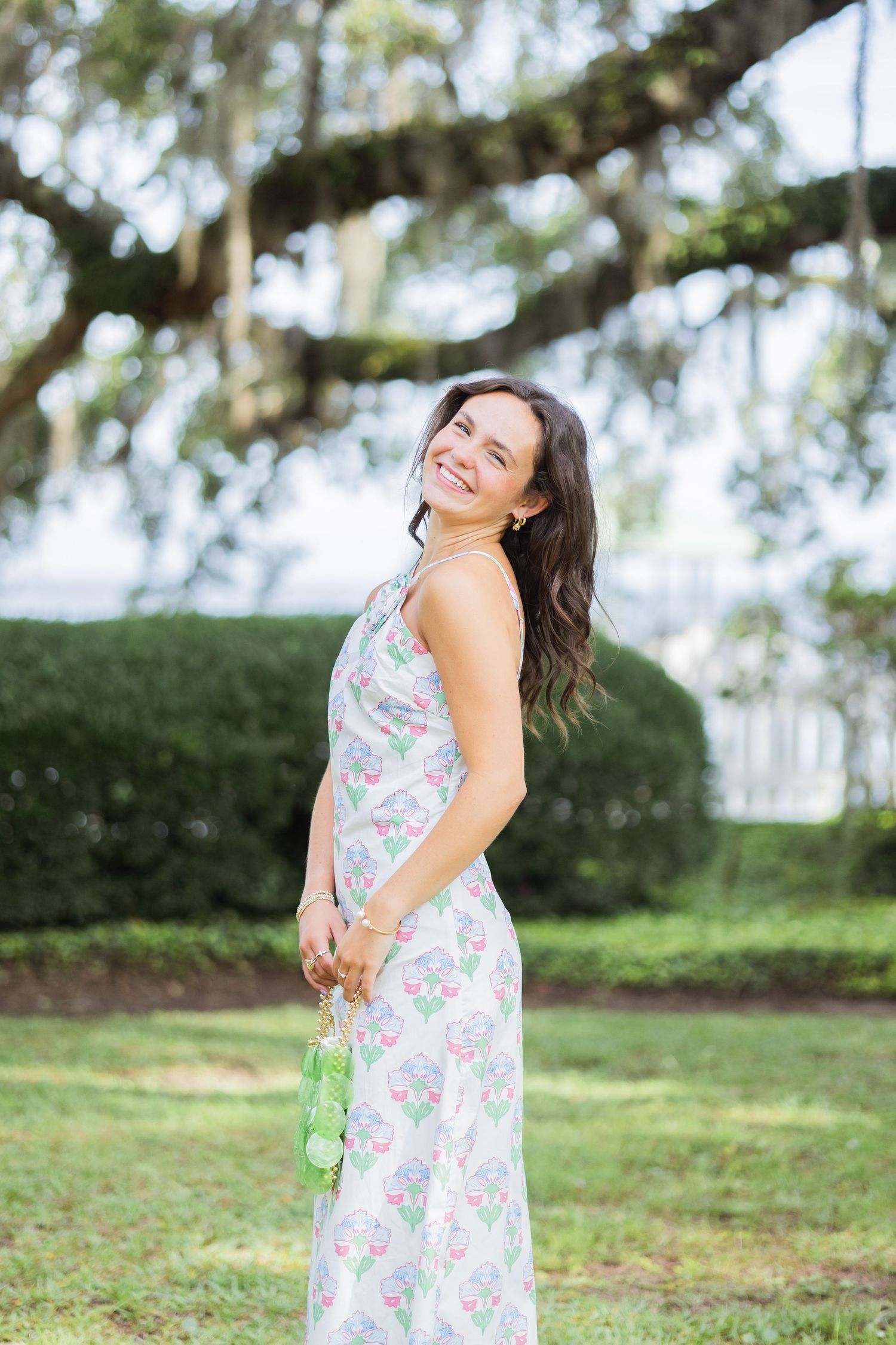 Smiling brunette woman in floral maxi dress holding green bag outdoors in lush garden.