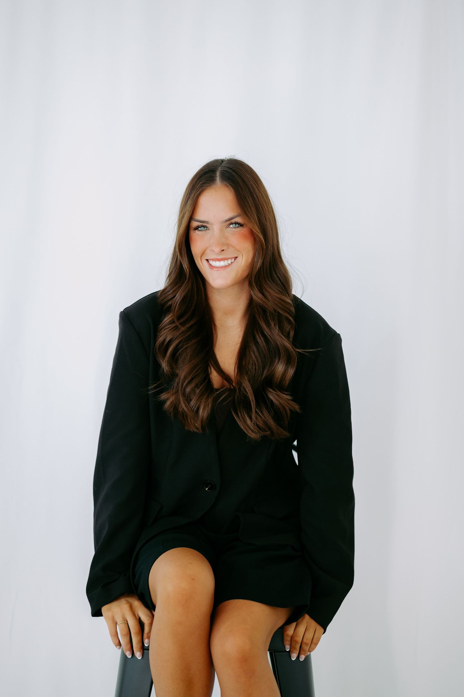 Smiling brunette woman in oversized black blazer dress seated on stool against white background.
