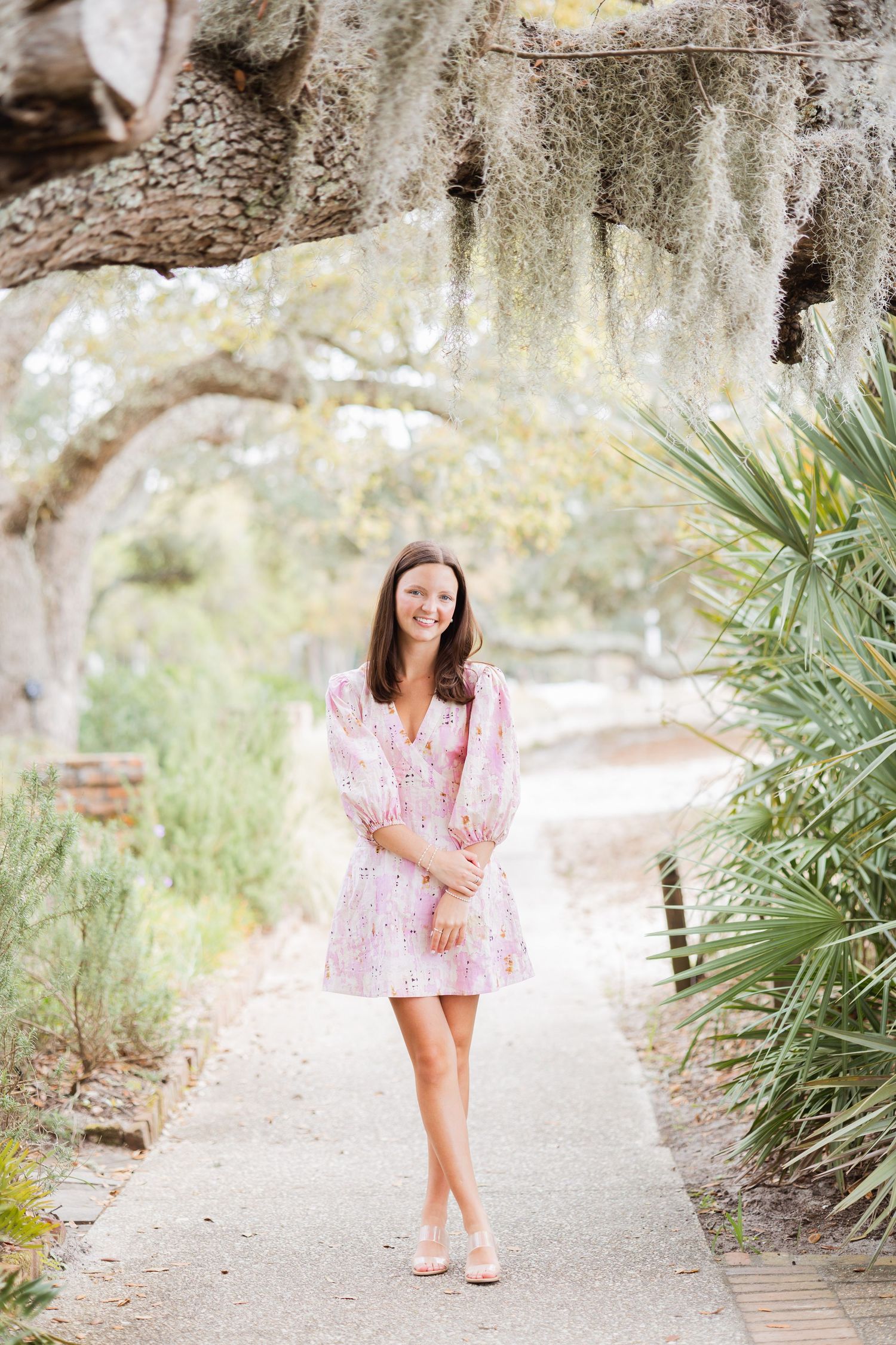 Brunette woman in pink floral dress smiling on a scenic garden path with Spanish moss and palm trees.