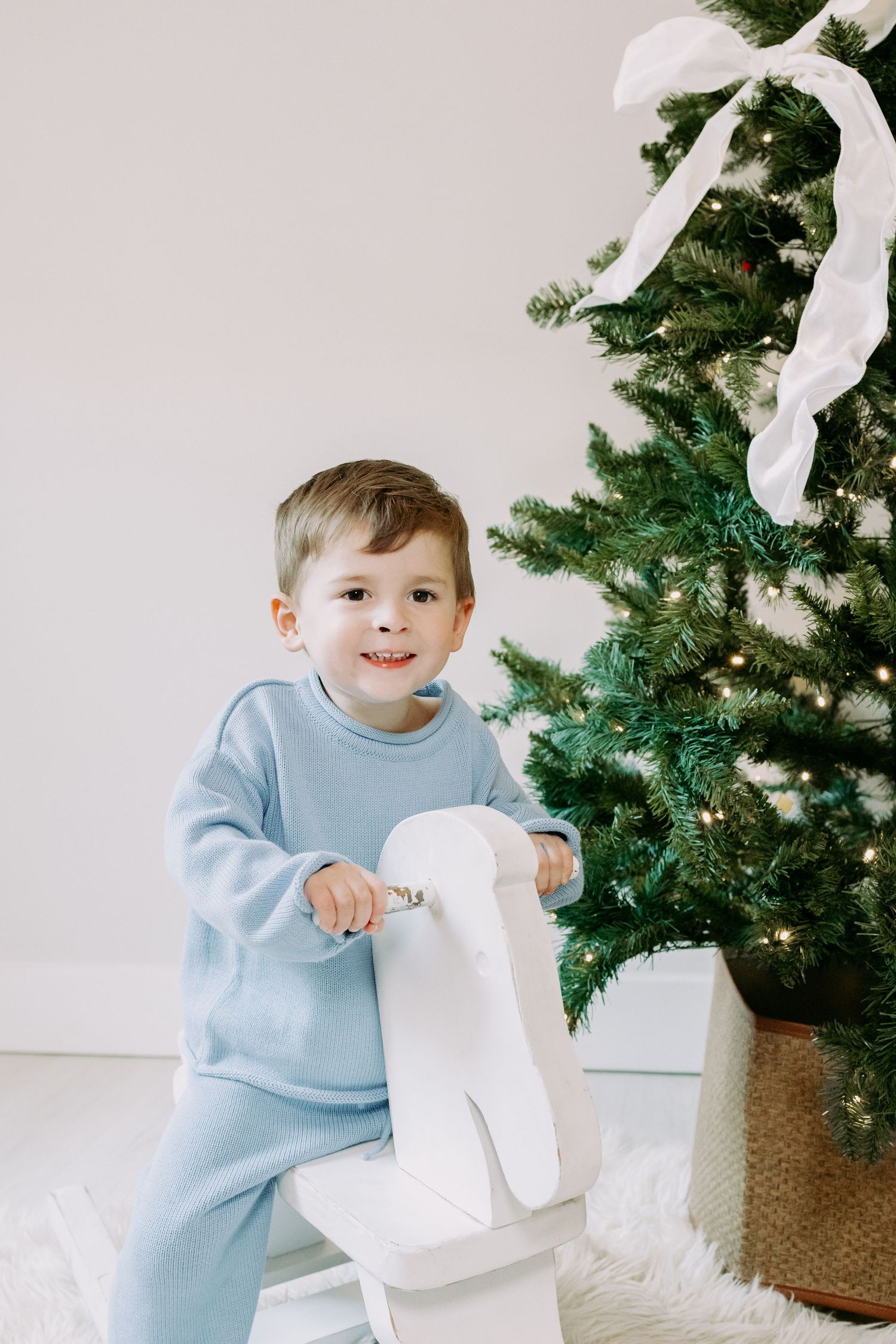Smiling toddler boy in a blue outfit sitting on a white rocking horse beside a decorated Christmas tree.