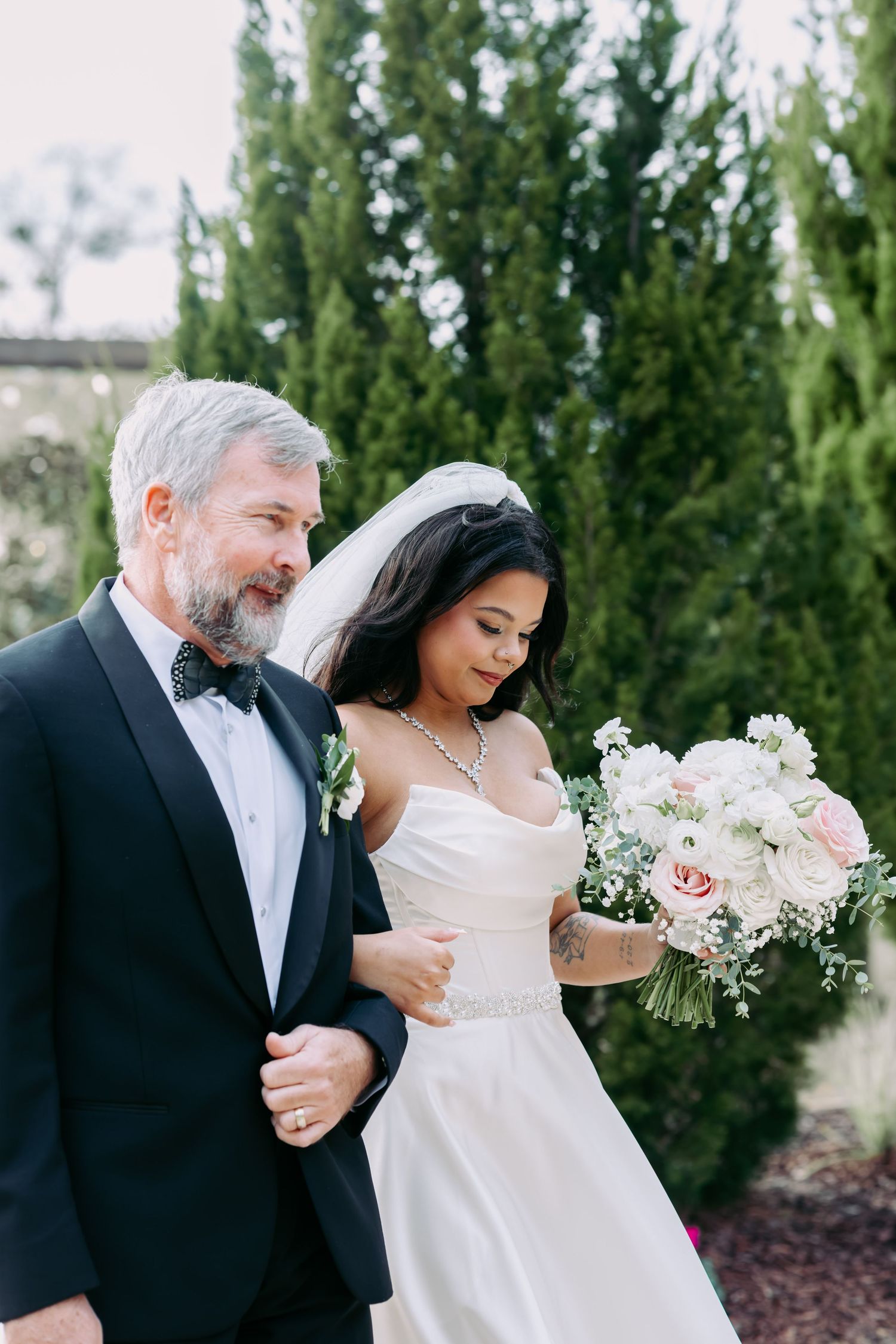 Father of the bride escorts his daughter in a strapless gown holding a pink rose bouquet.