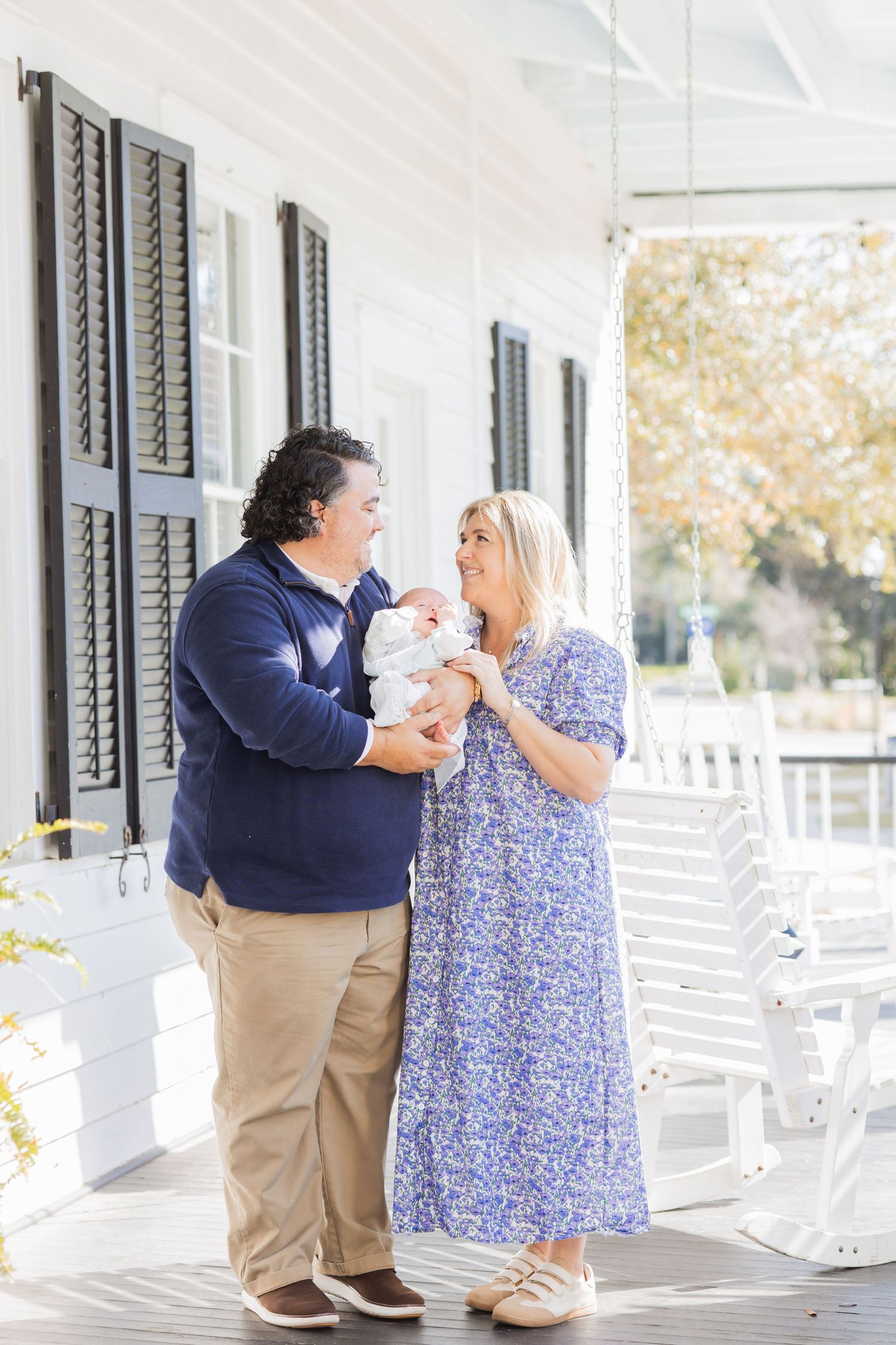 Couple in coordinating blue outfits smiling at their newborn baby on a white Southern porch.