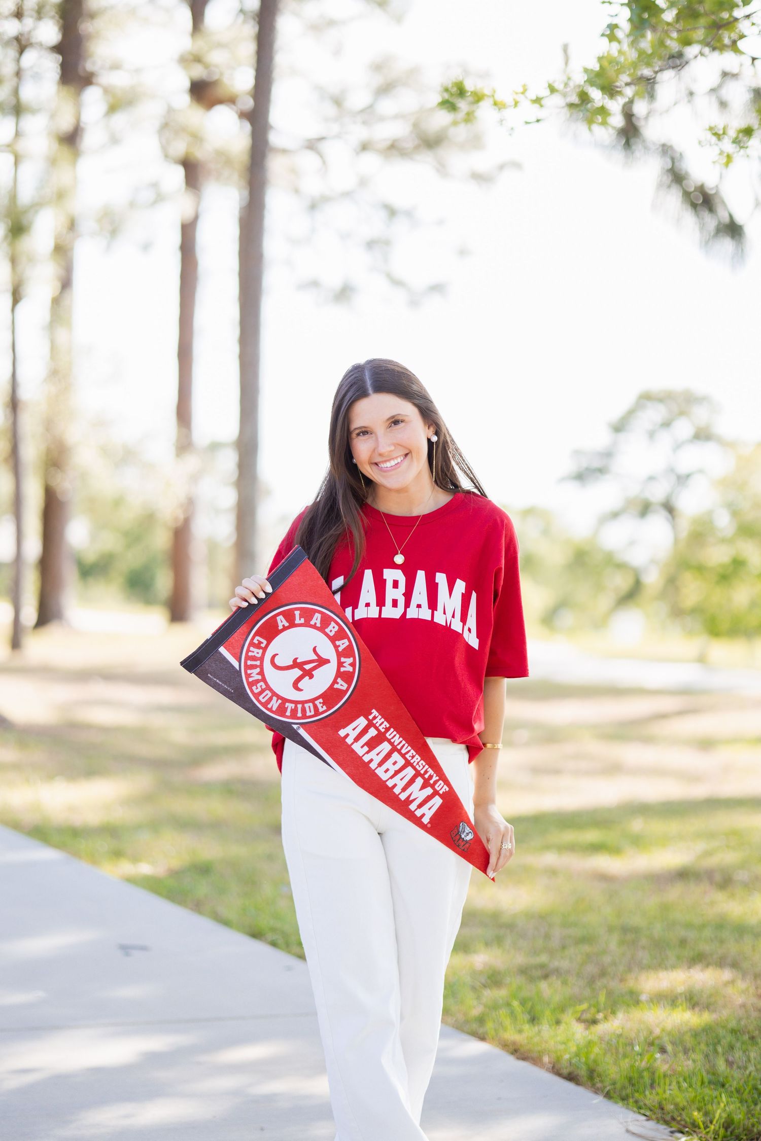 Smiling young woman holding University of Alabama pennant and wearing red Alabama t-shirt outdoors.