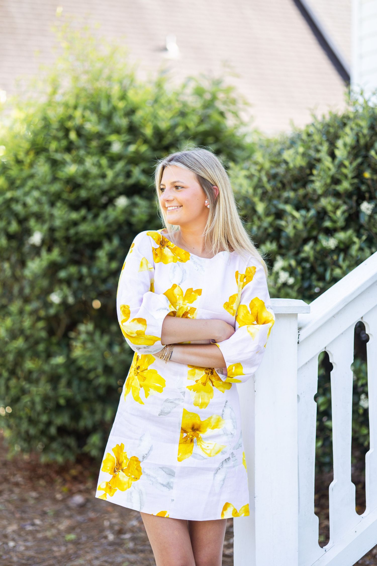 Blonde woman in a yellow floral dress smiling while leaning on a white railing outdoors.