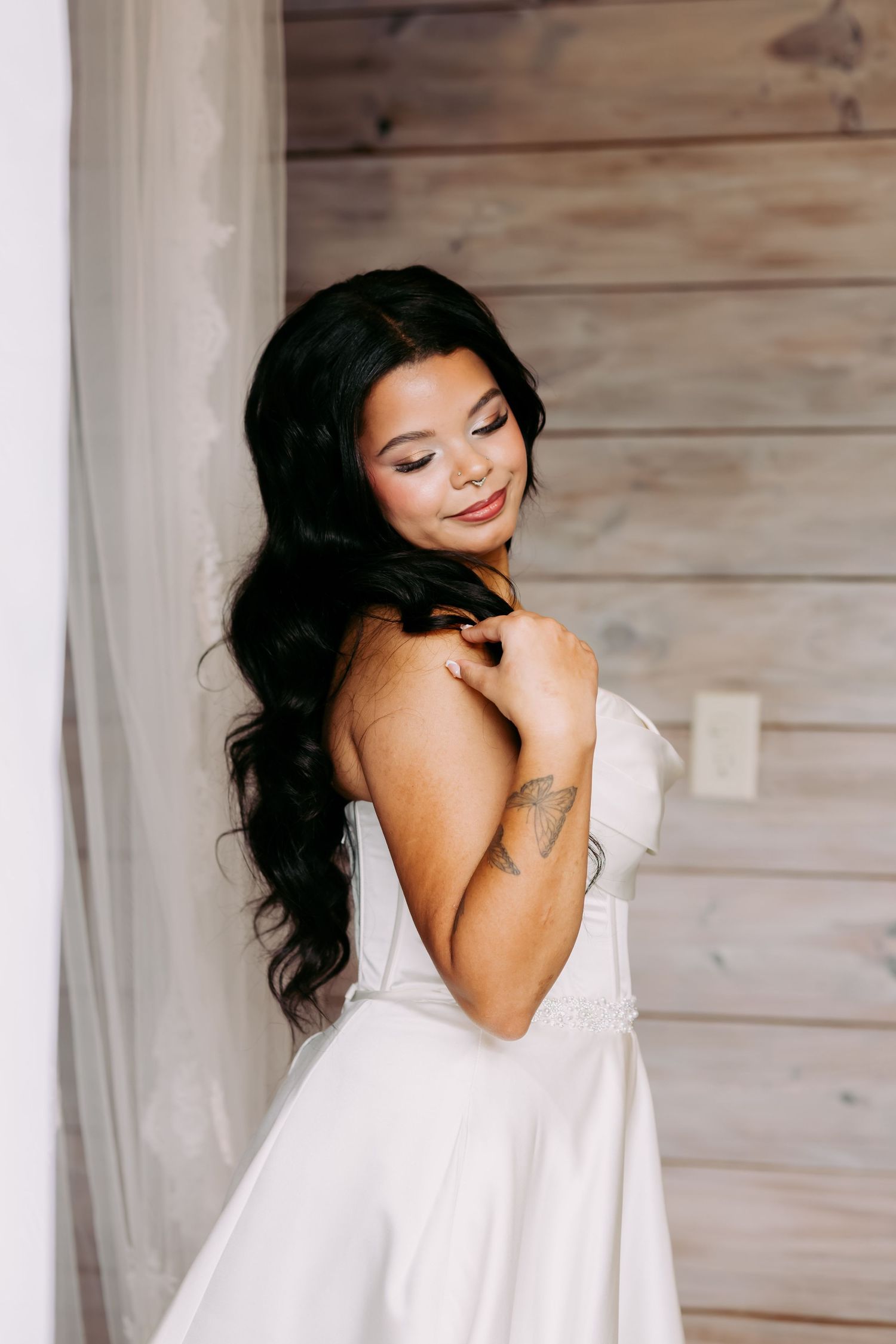 Beautiful dark-haired bride in strapless white satin gown posing gracefully against rustic wood-paneled wall.