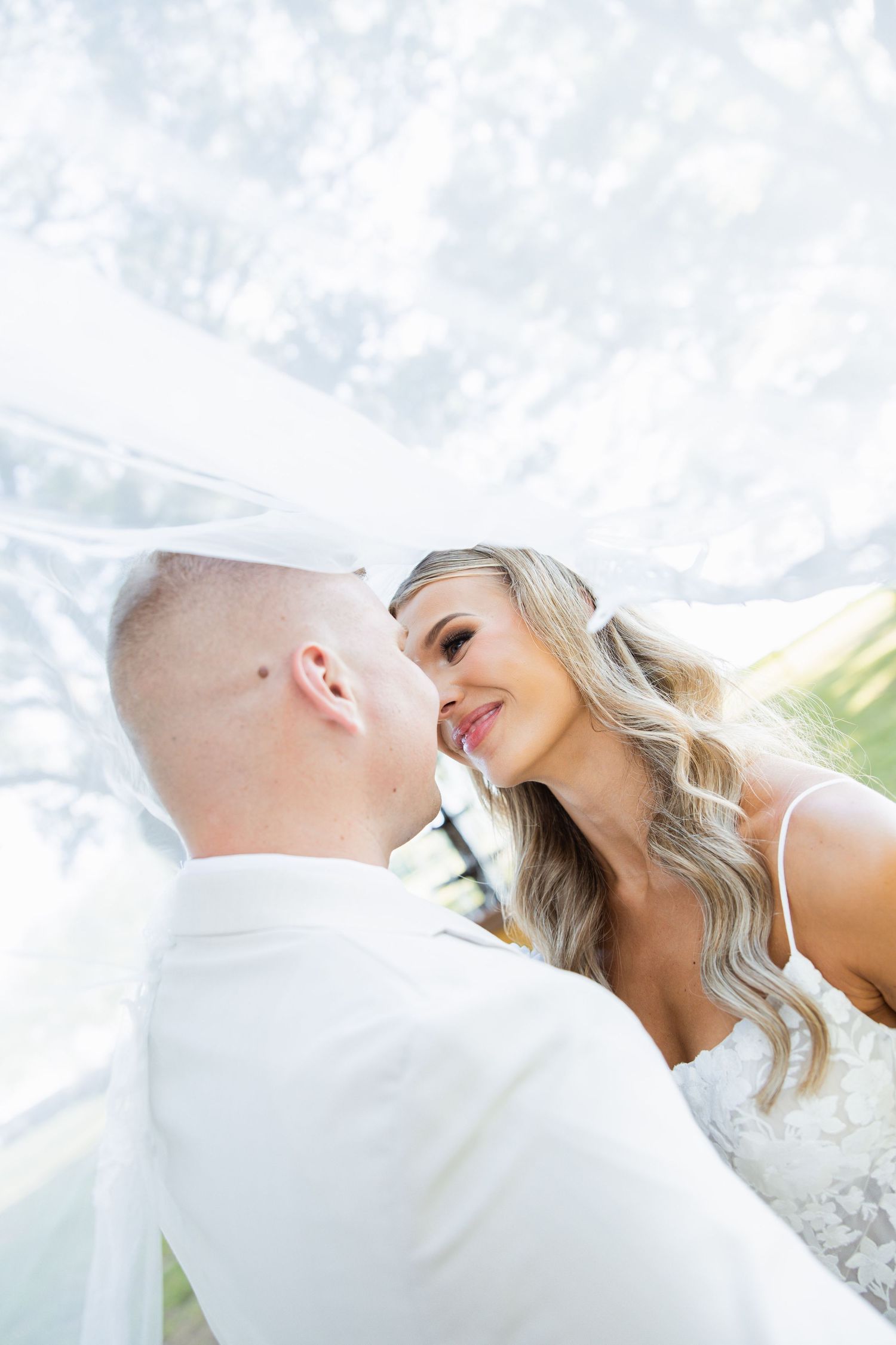 Romantic bride and groom share intimate moment under flowing white veil outdoors on wedding day.
