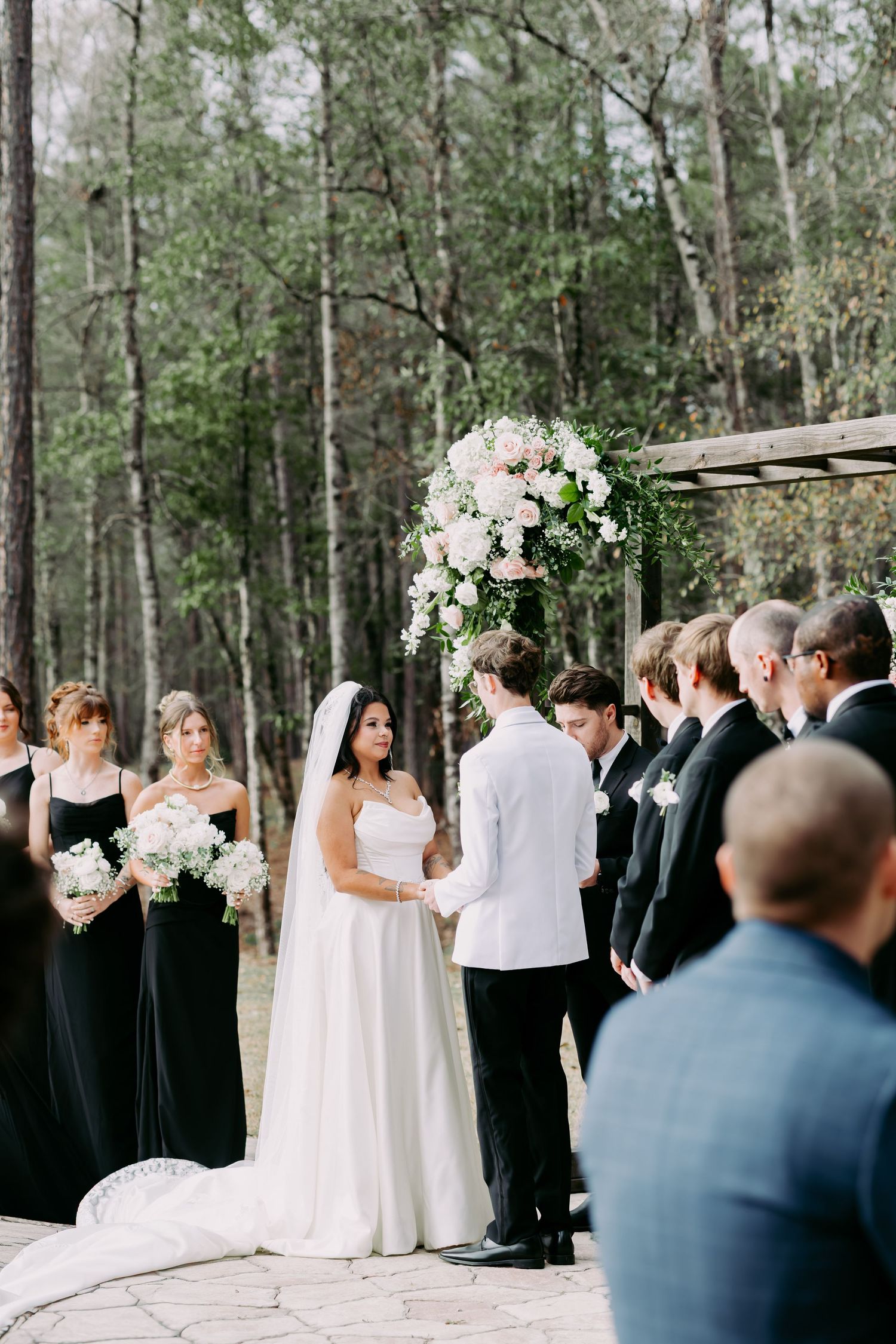 Outdoor wedding ceremony in forest with bride and groom at floral arch, bridesmaids in black gowns.