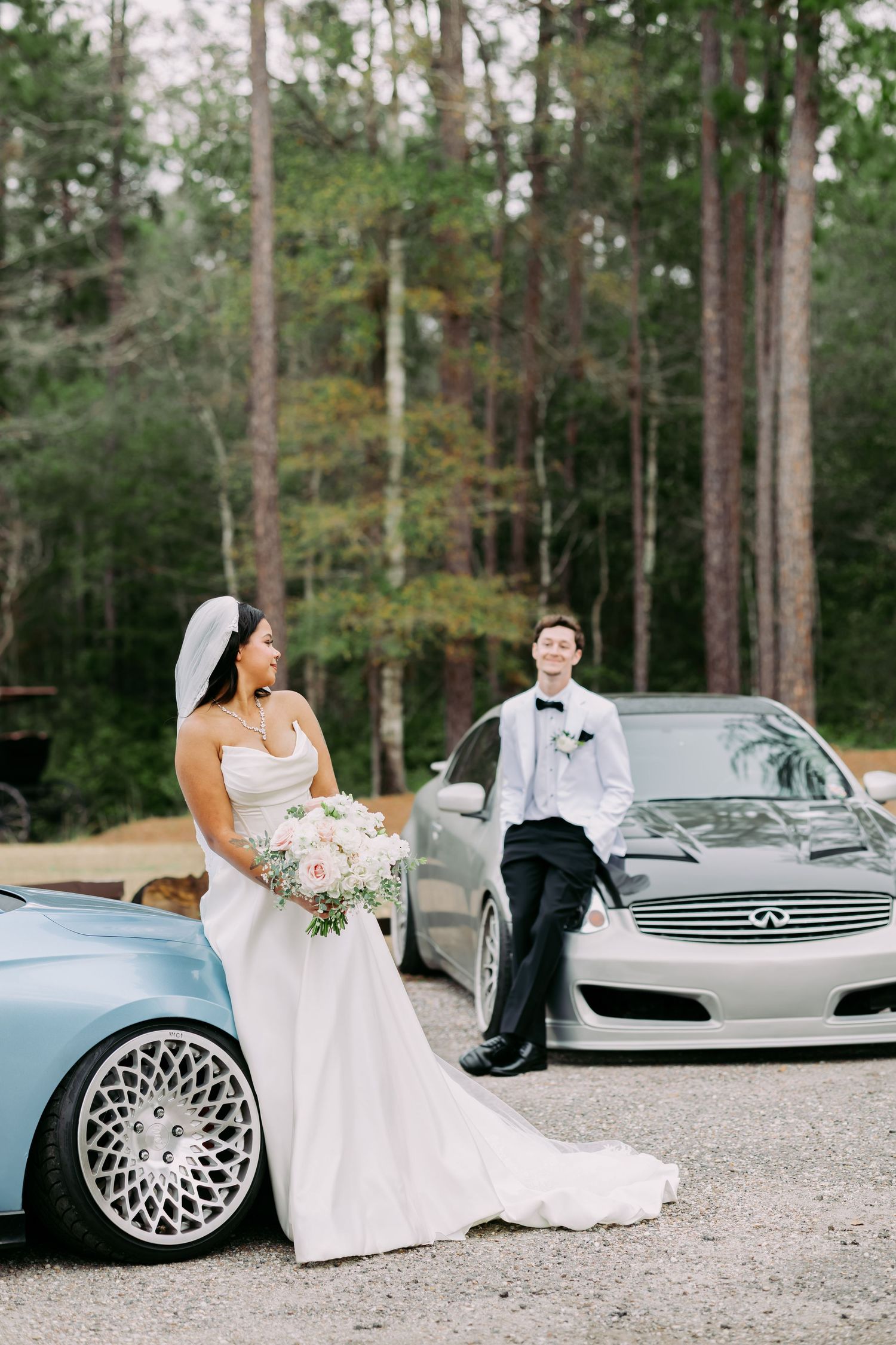Bride and groom posing with two custom cars in a wooded setting on their wedding day.