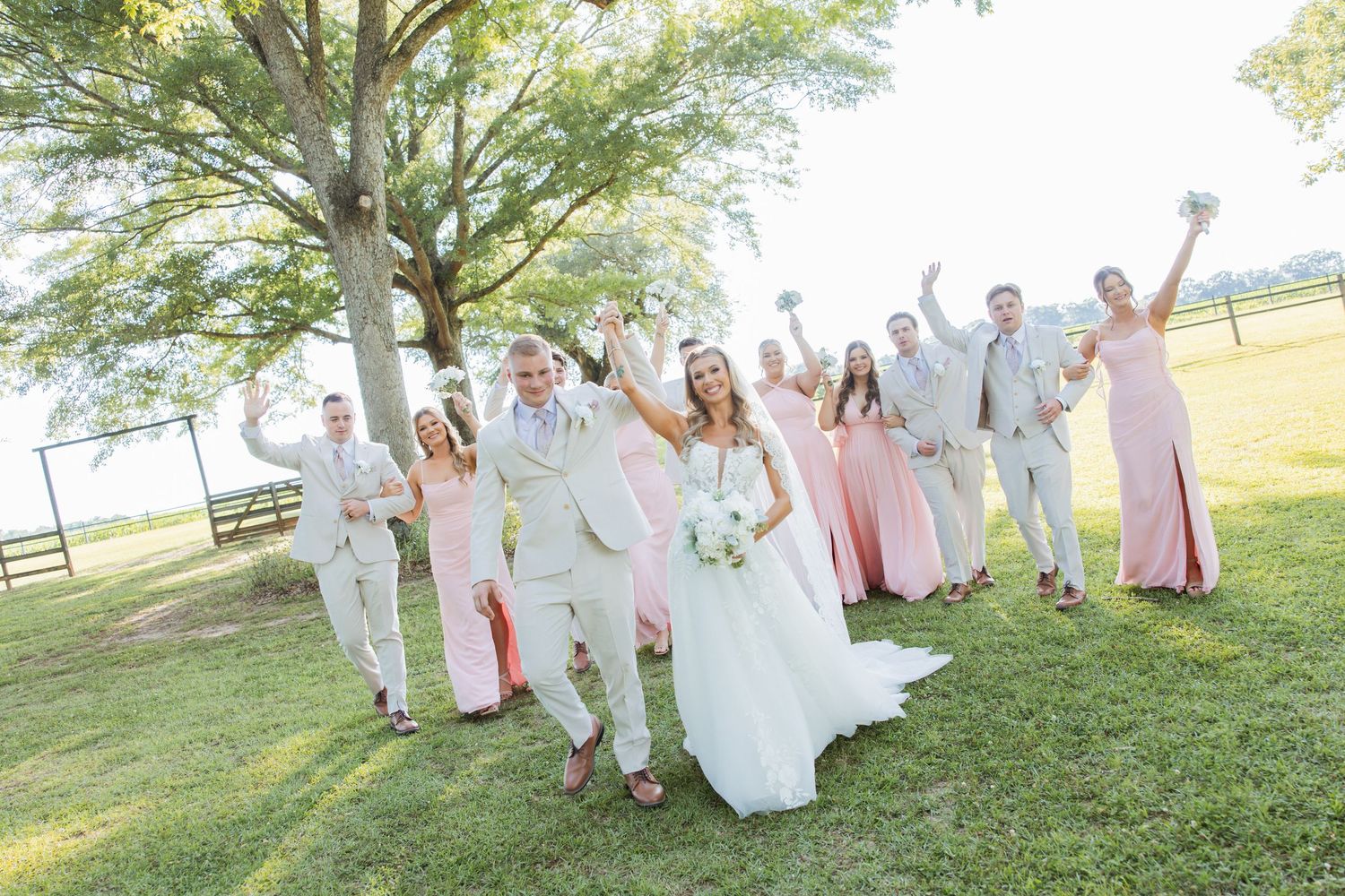 Joyful wedding party celebrates on green lawn with bridesmaids in pink dresses and groomsmen in tan suits.