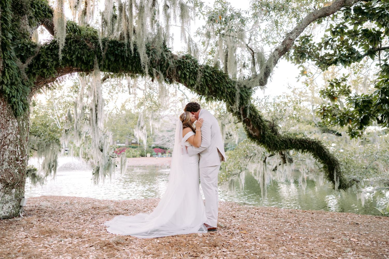 Bride and groom kiss under sweeping Spanish moss-covered oak trees beside a serene pond at outdoor wedding.