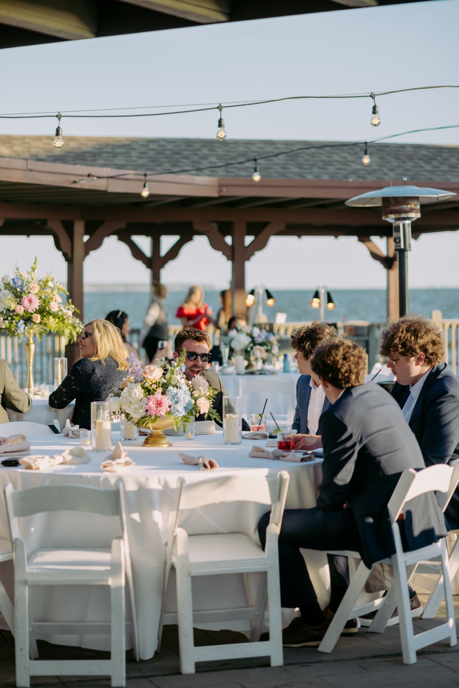 Guests dining at outdoor waterfront wedding reception with string lights and colorful floral centerpieces.