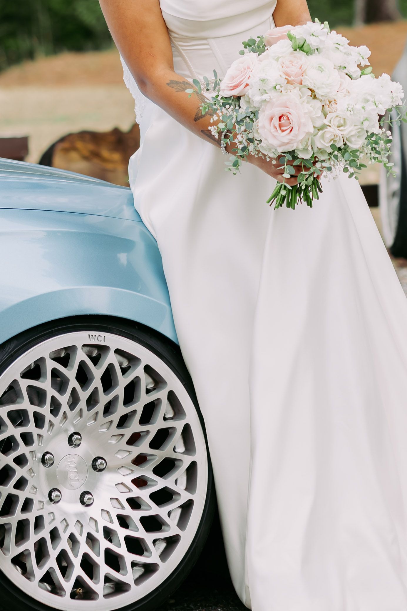 Bride in white gown holding pink rose bouquet beside a baby blue car with ornate white wheels.