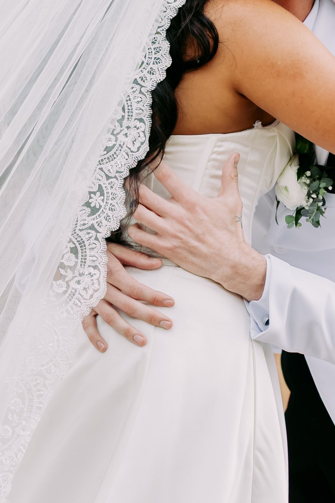 Close-up of groom's hands embracing bride's waist, showcasing lace-trimmed cathedral veil detail.