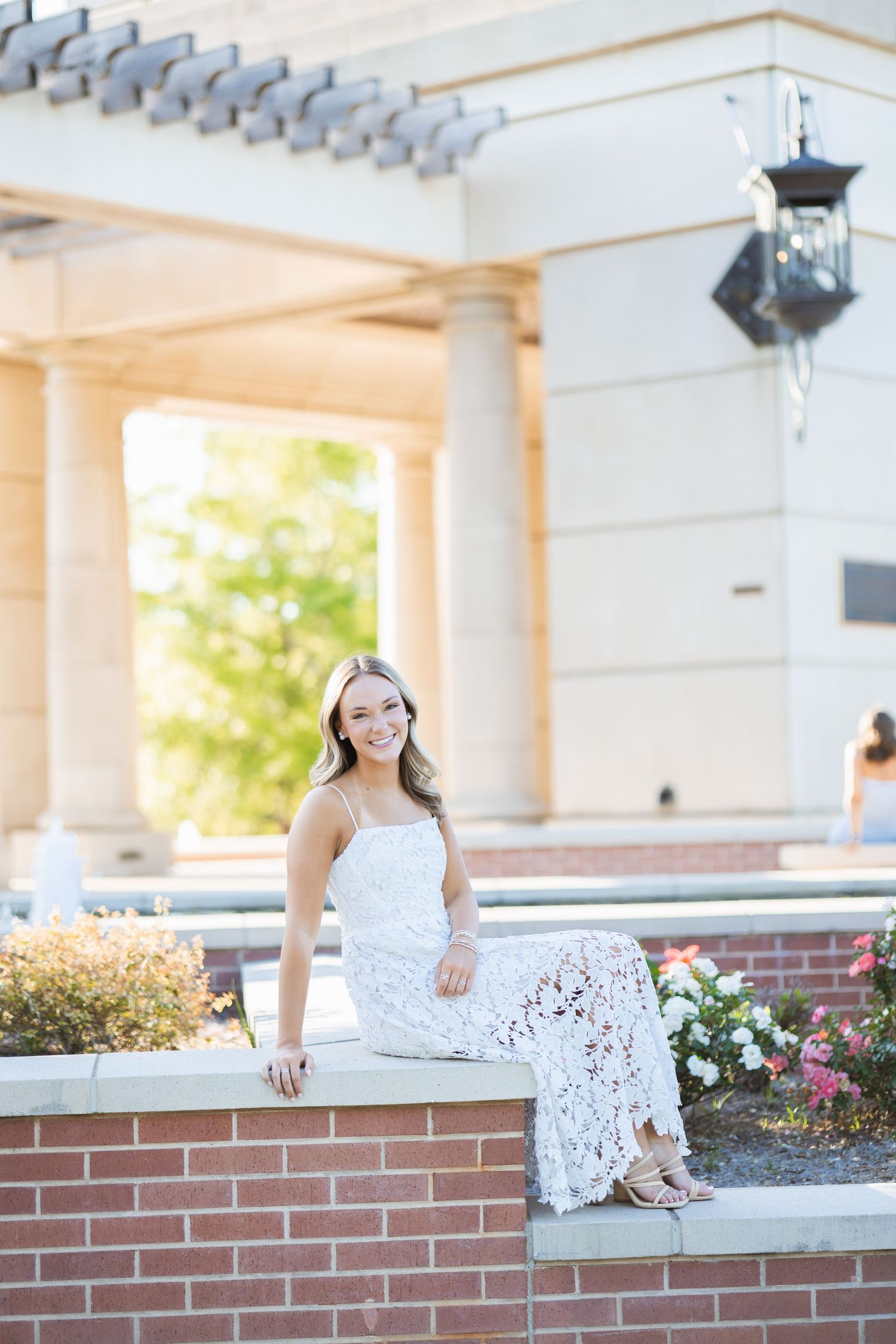 Smiling young woman in a white lace dress sitting on a brick wall near a columned building.