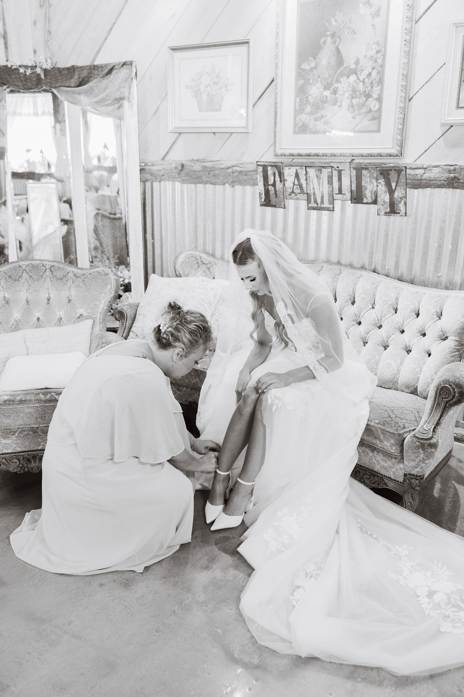 Black and white photo of bridesmaid helping bride put on wedding shoes while seated on elegant tufted sofa.