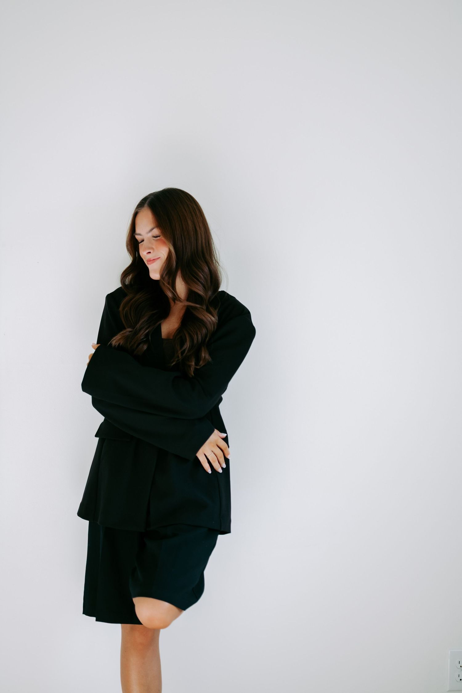 Brunette woman in black blazer and shorts standing with arms crossed smiling against white wall.