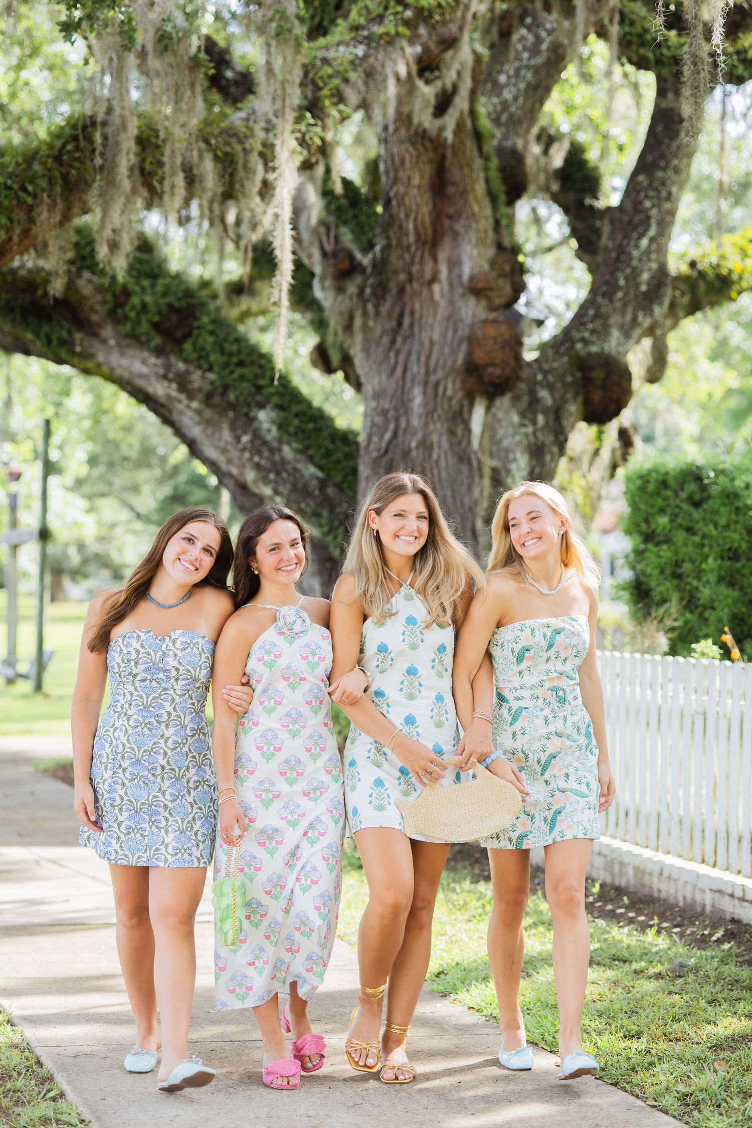 Four smiling women in pastel summer dresses walking together under a large oak tree.