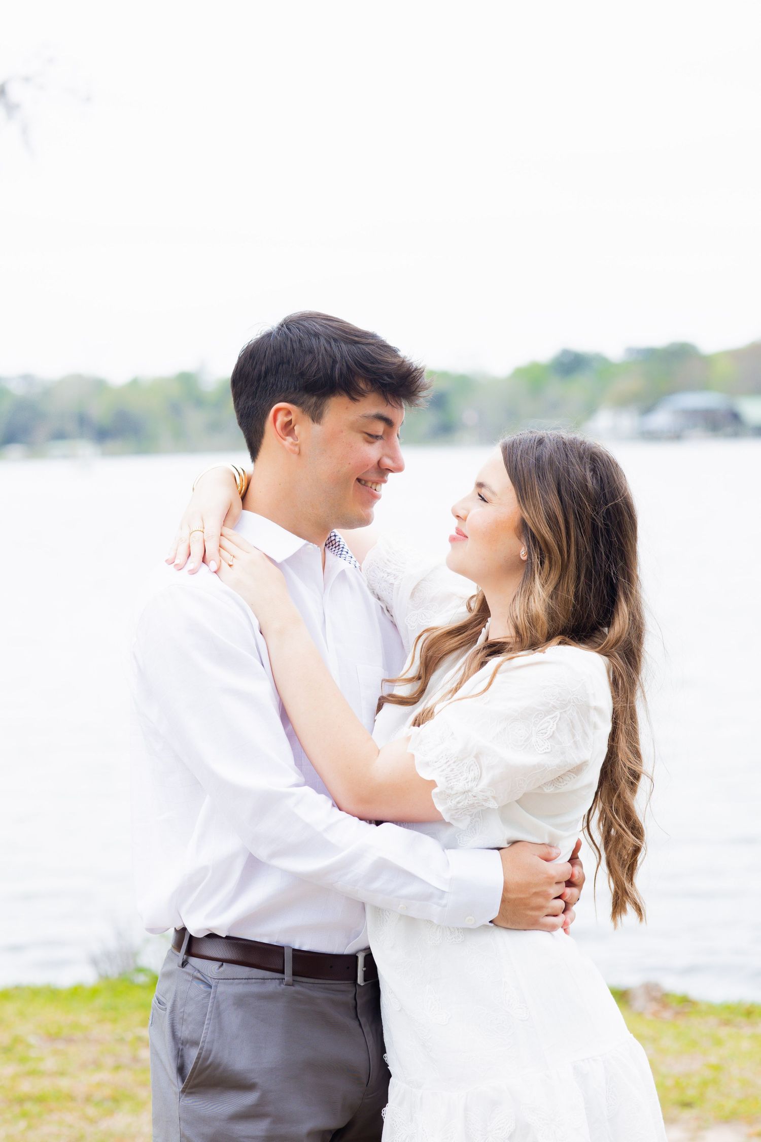 Young couple embracing and smiling at each other near a sunlit waterfront backdrop.