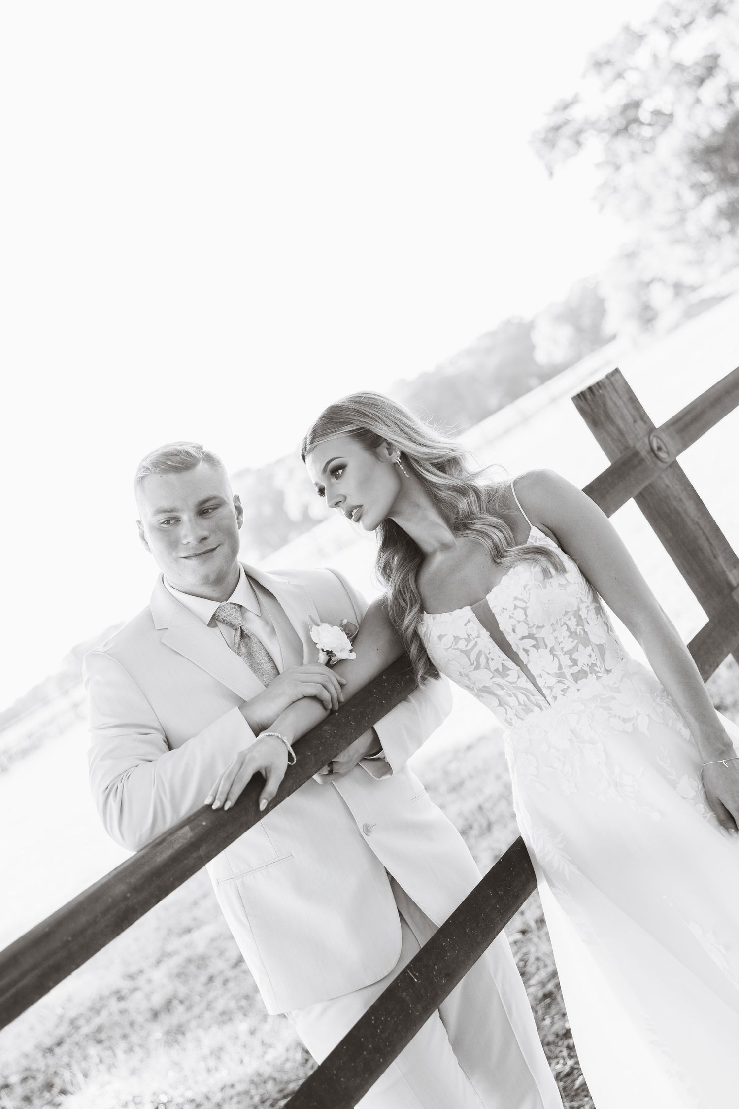 Black and white photo of bride and groom leaning on a wooden fence at their outdoor wedding.