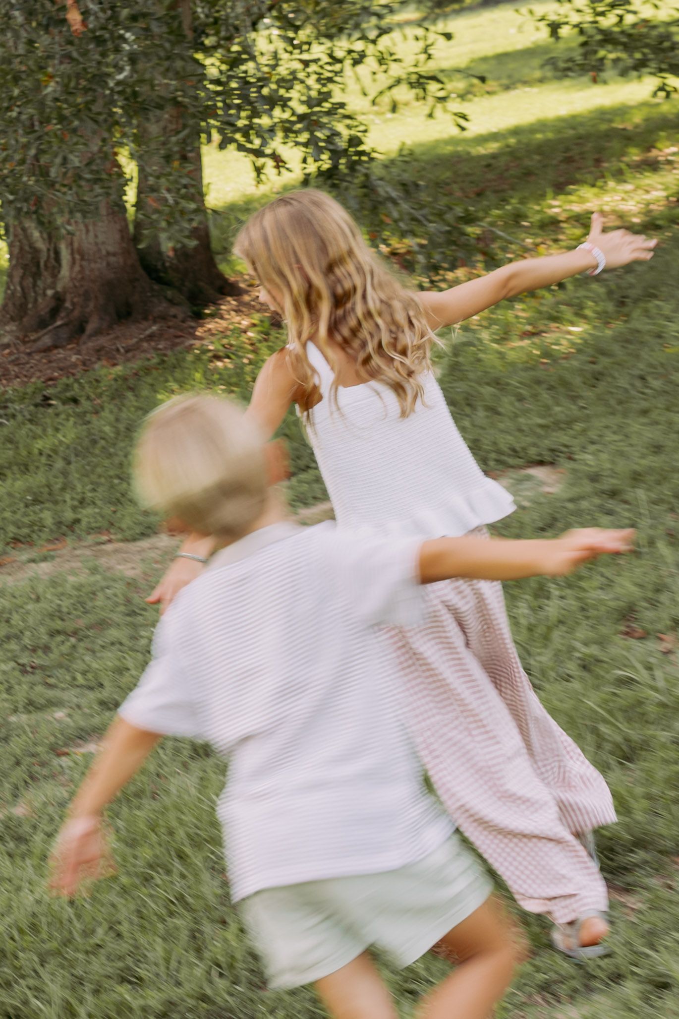 Two children running playfully on green grass near a large tree in a park.