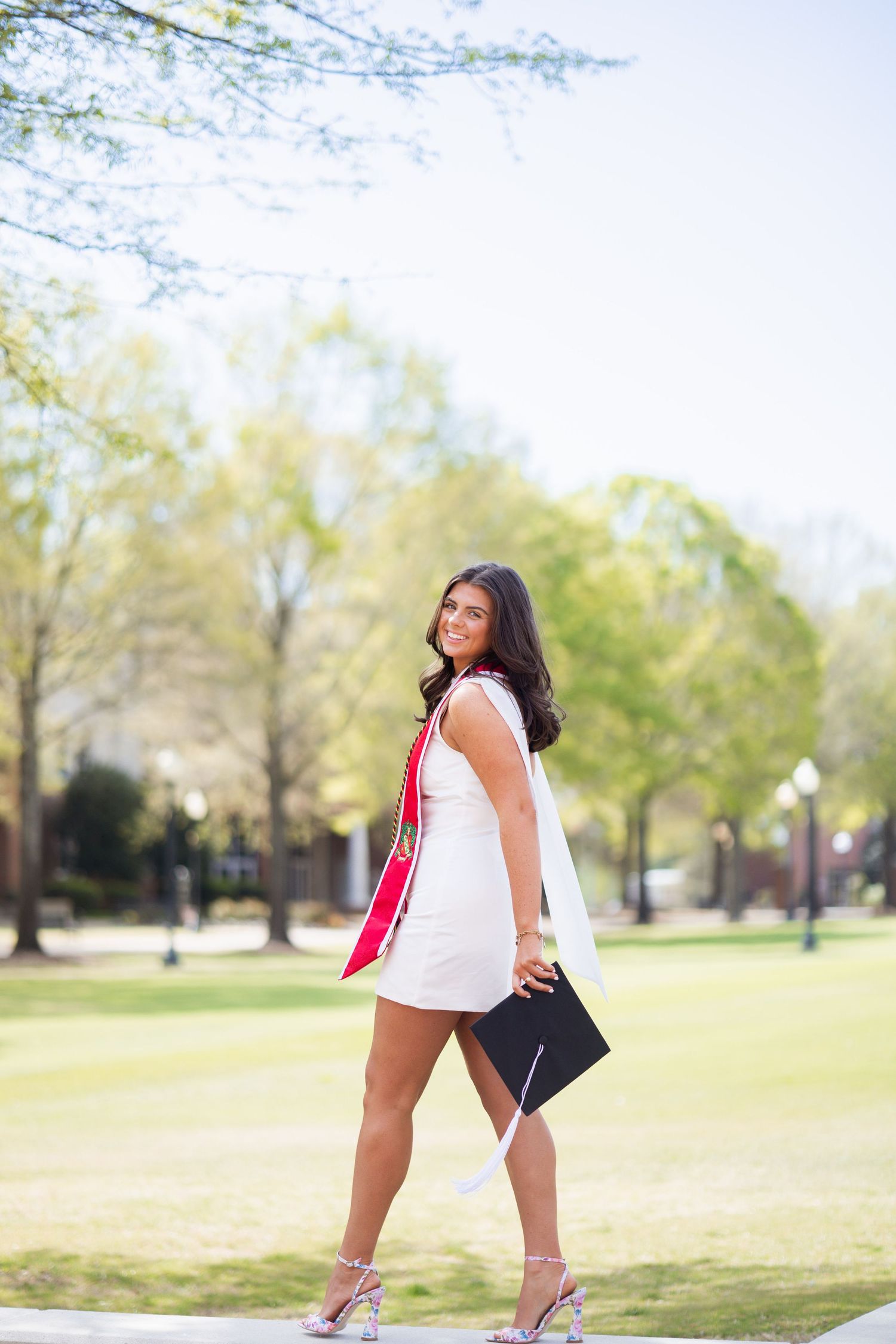Graduating woman in white dress holding cap and red sash, walking on a tree-lined campus path.