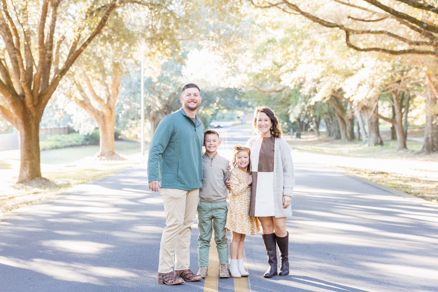 A family of four stands together on a tree-lined road, smiling in coordinating neutral fall outfits.