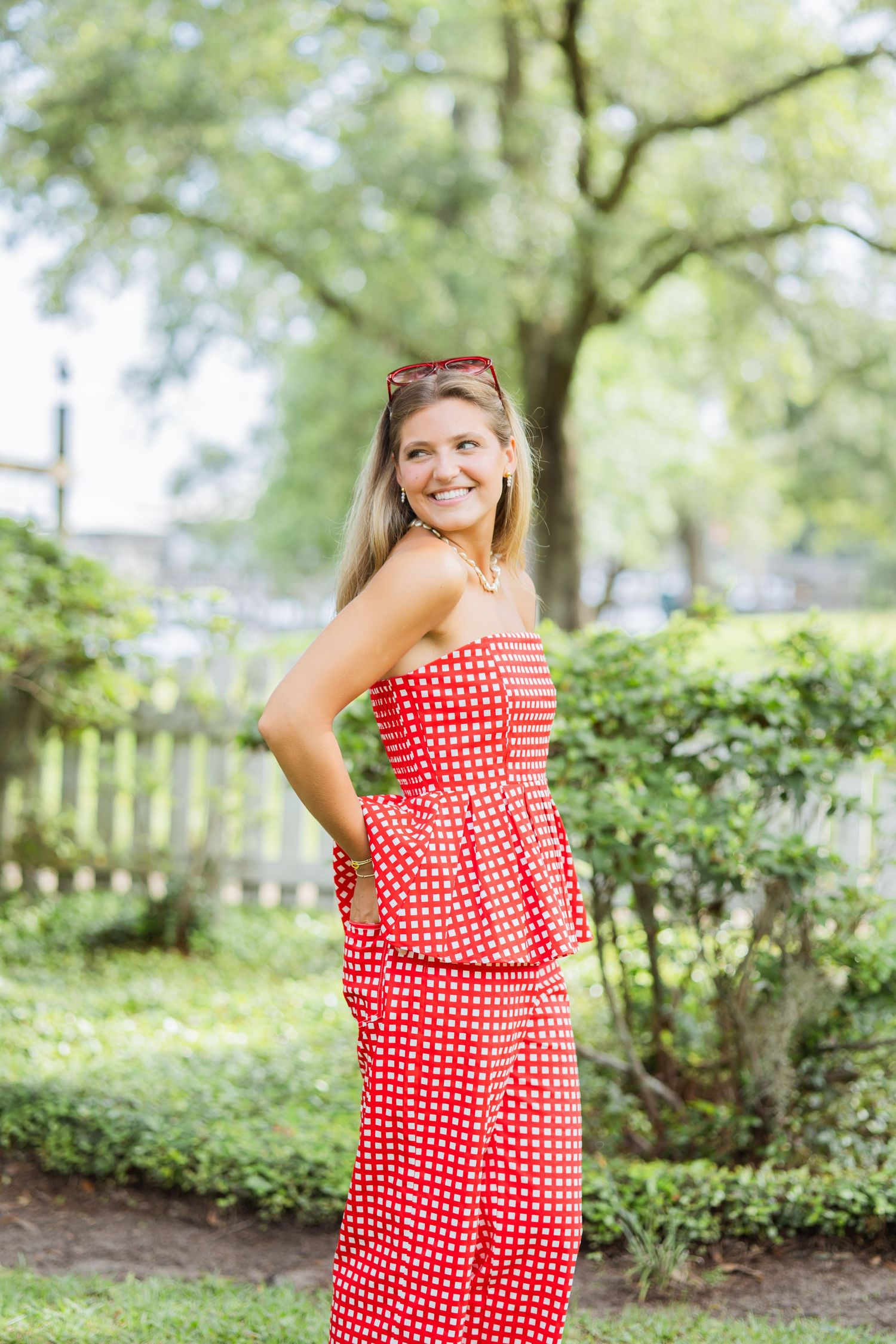 Smiling blonde woman in red polka dot strapless jumpsuit posing in lush garden setting.