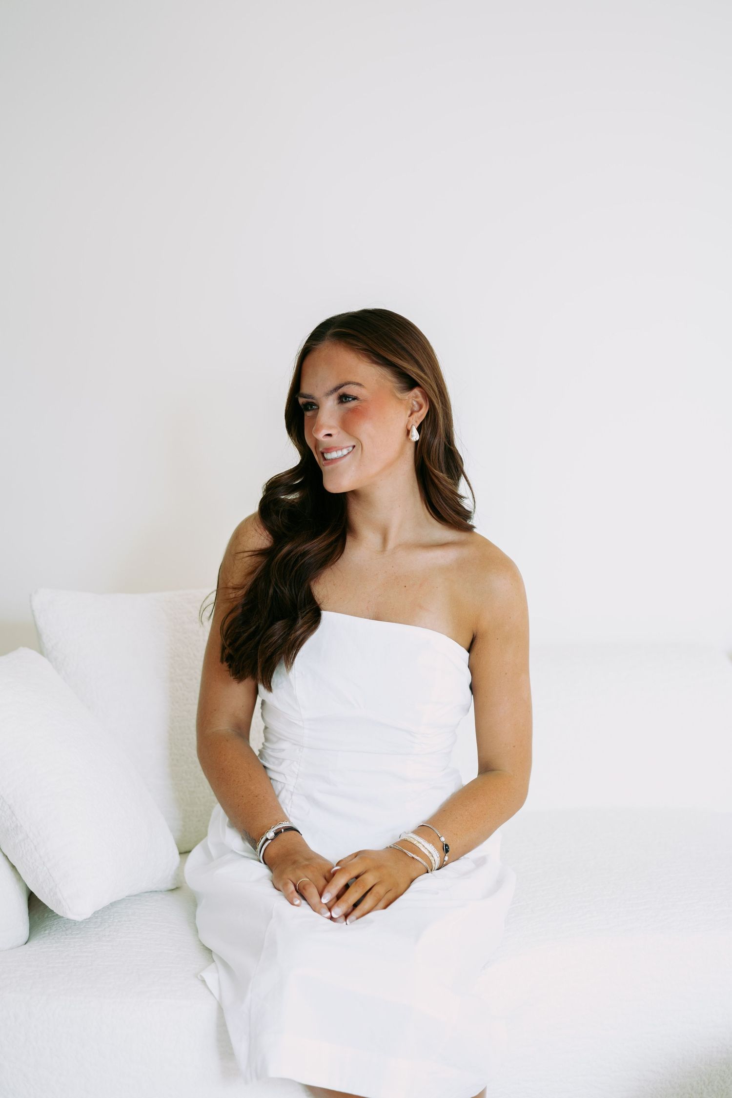 Smiling brunette woman in white strapless dress seated on white sofa wearing silver bracelets.
