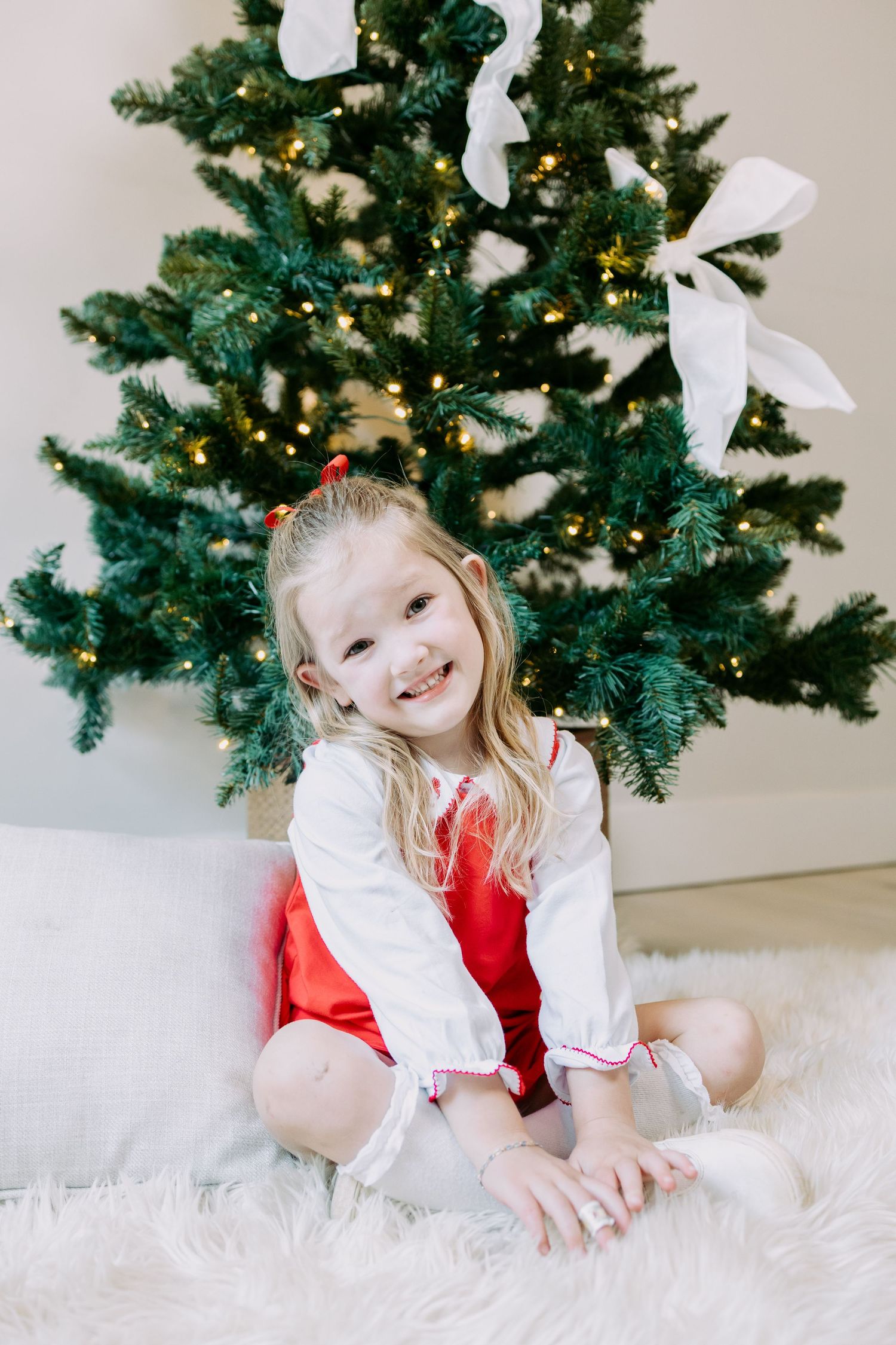 Happy little girl in a red and white outfit sitting on a fluffy rug in front of a lit Christmas tree.