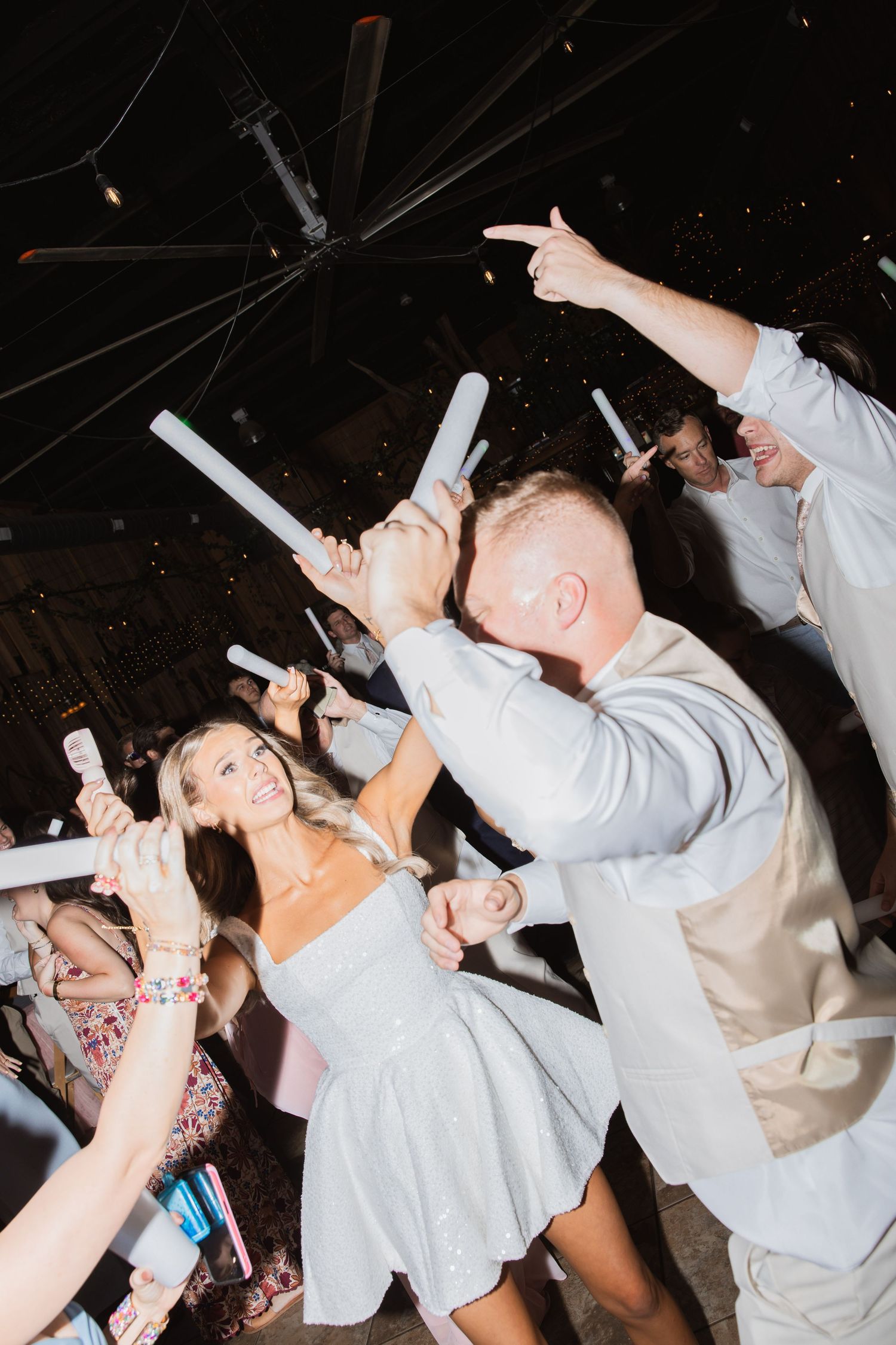 Bride and groom dance energetically with guests holding glow sticks at lively wedding reception.