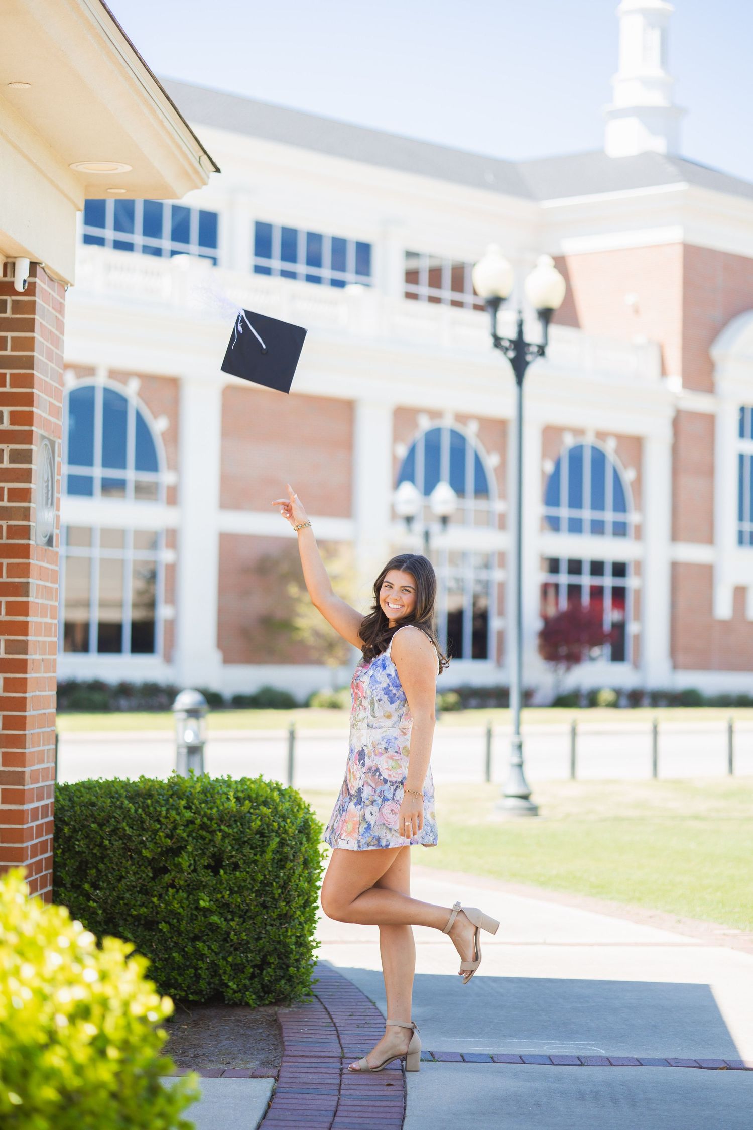 Smiling graduate in floral dress tossing cap in front of brick university building.