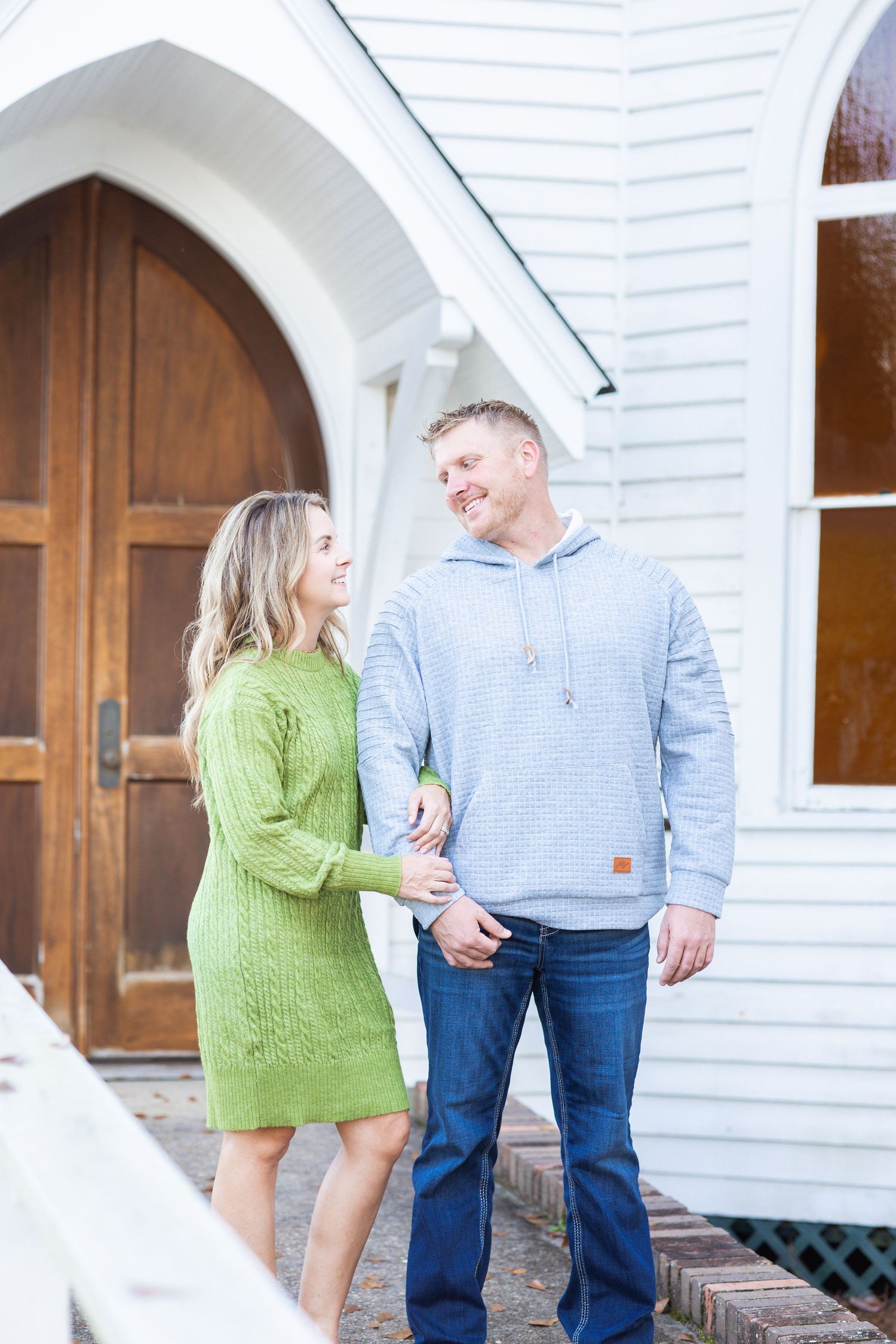Smiling couple holding hands in front of a white chapel with wooden arched doors in casual outfits.