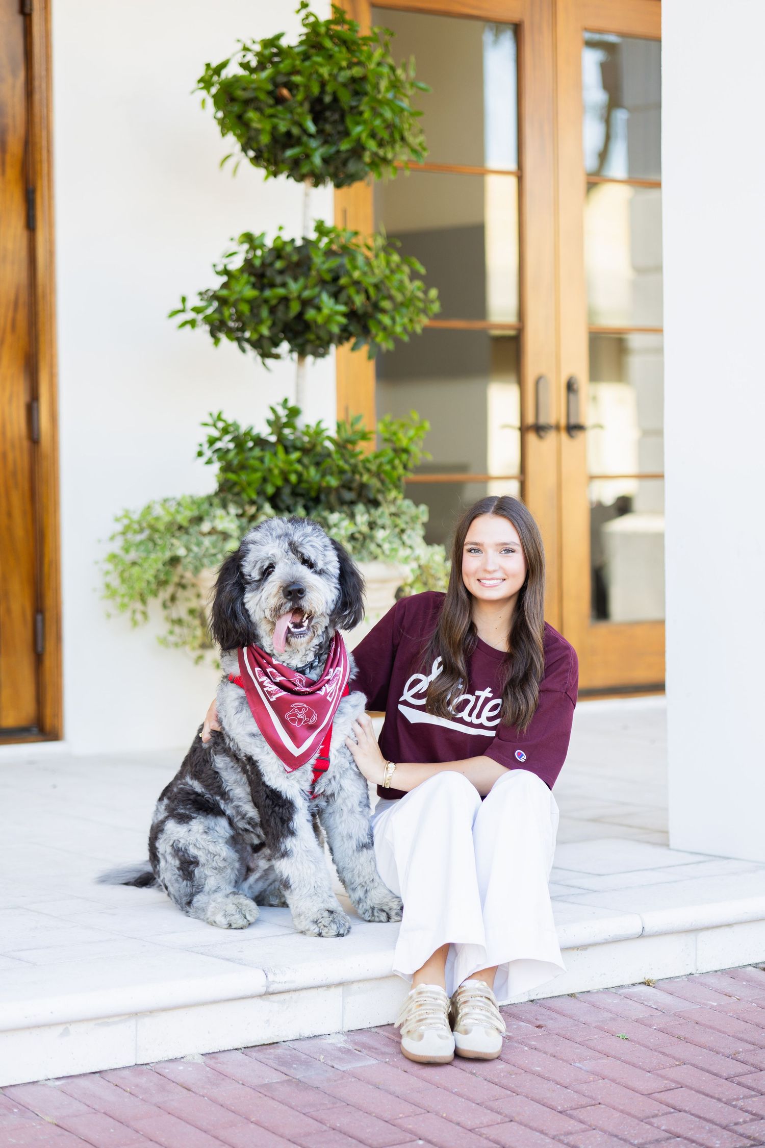 Smiling young woman sitting with large fluffy dog wearing red bandana on home entrance steps.