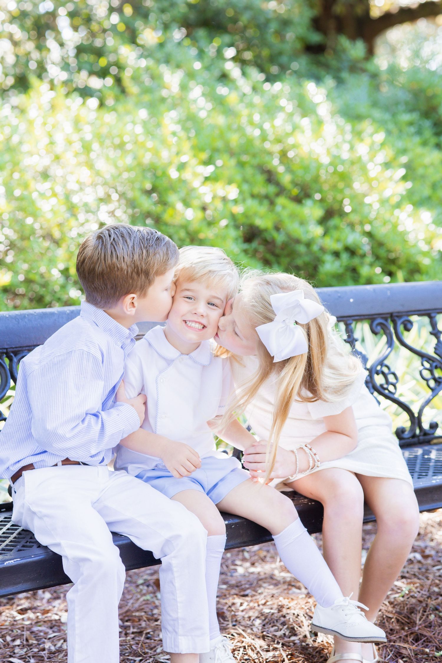 Two siblings give their younger brother a kiss on the cheeks while sitting on a decorative bench in a sunny park.