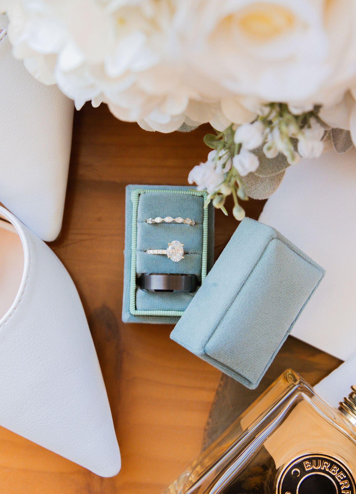 Close-up of diamond engagement ring and wedding bands in teal velvet ring box with white florals.