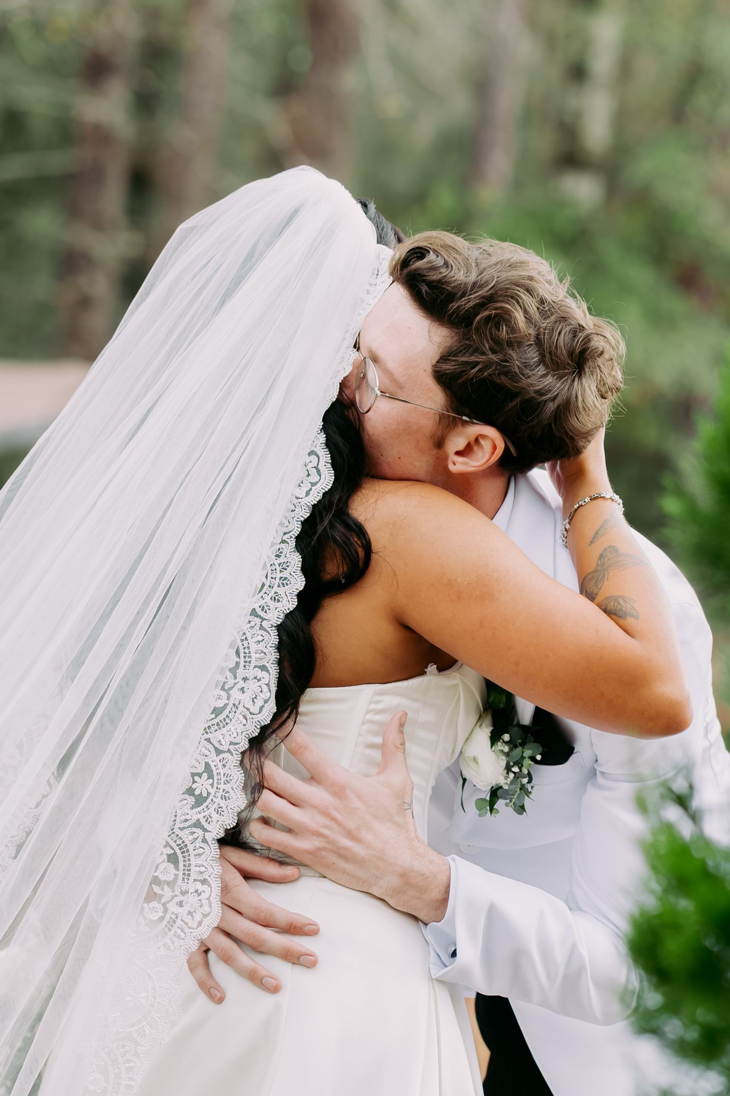 Bride and groom share a romantic first kiss embrace, her lace-edged veil flowing, surrounded by lush green trees.