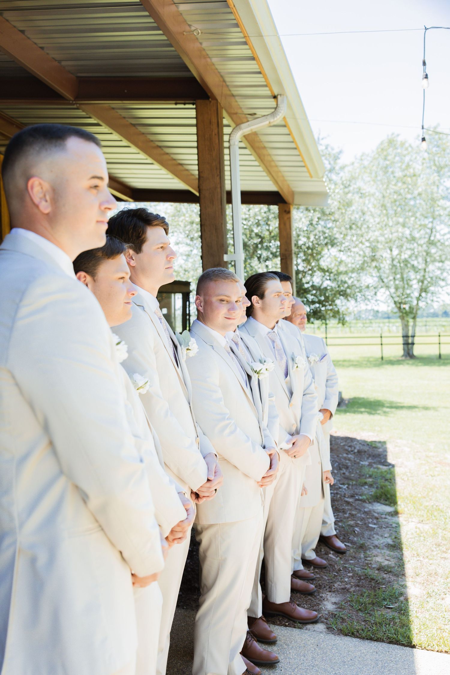 Groom and groomsmen in tan suits stand in a line under a covered porch outdoors.