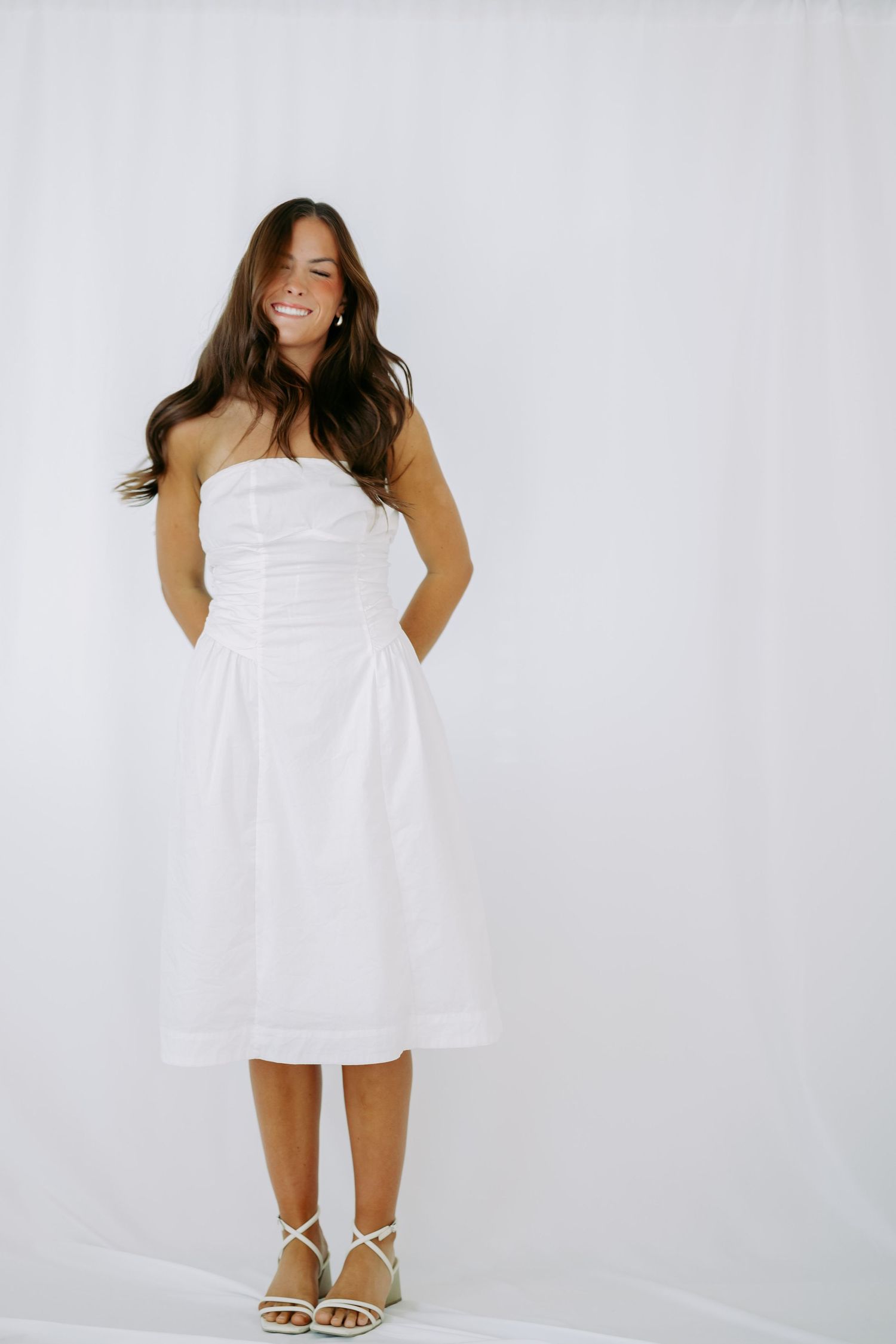 Woman in white strapless midi dress with ruched bodice standing and smiling against white backdrop.