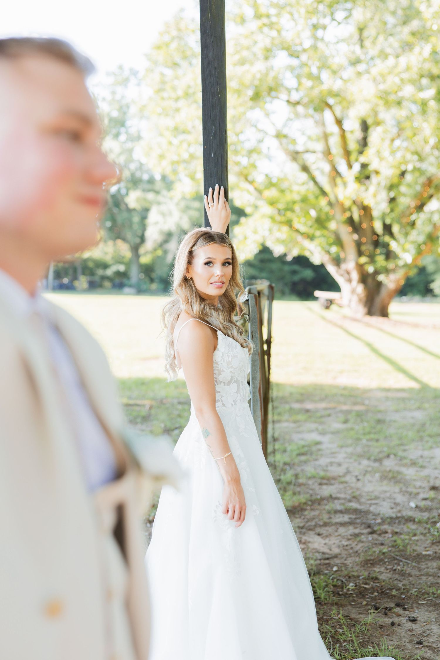 Bride in lace wedding gown stands by rustic barn door while groom stands in foreground.