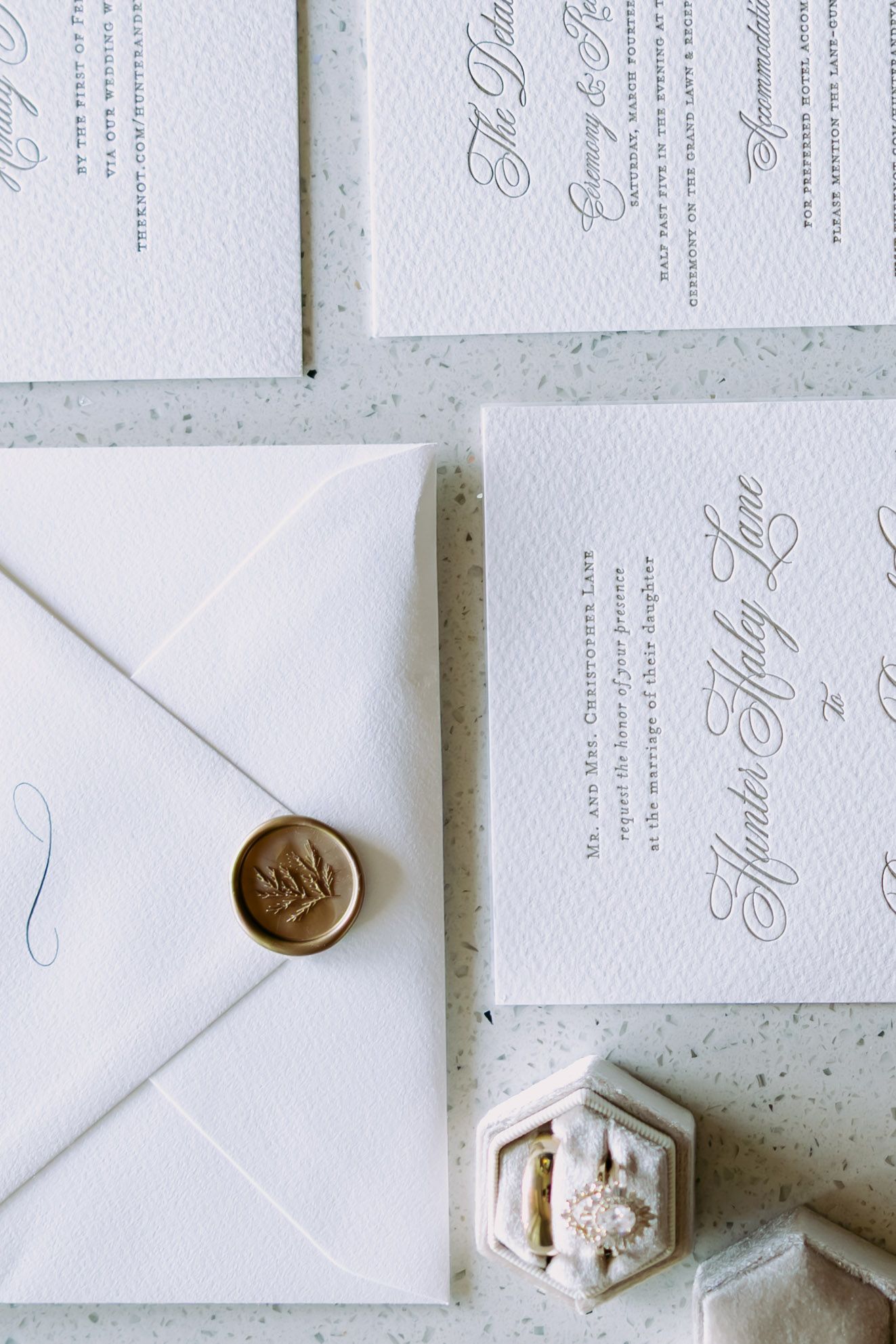 Elegant wedding invitation suite with gold wax seal envelope and velvet ring box on a marble surface.