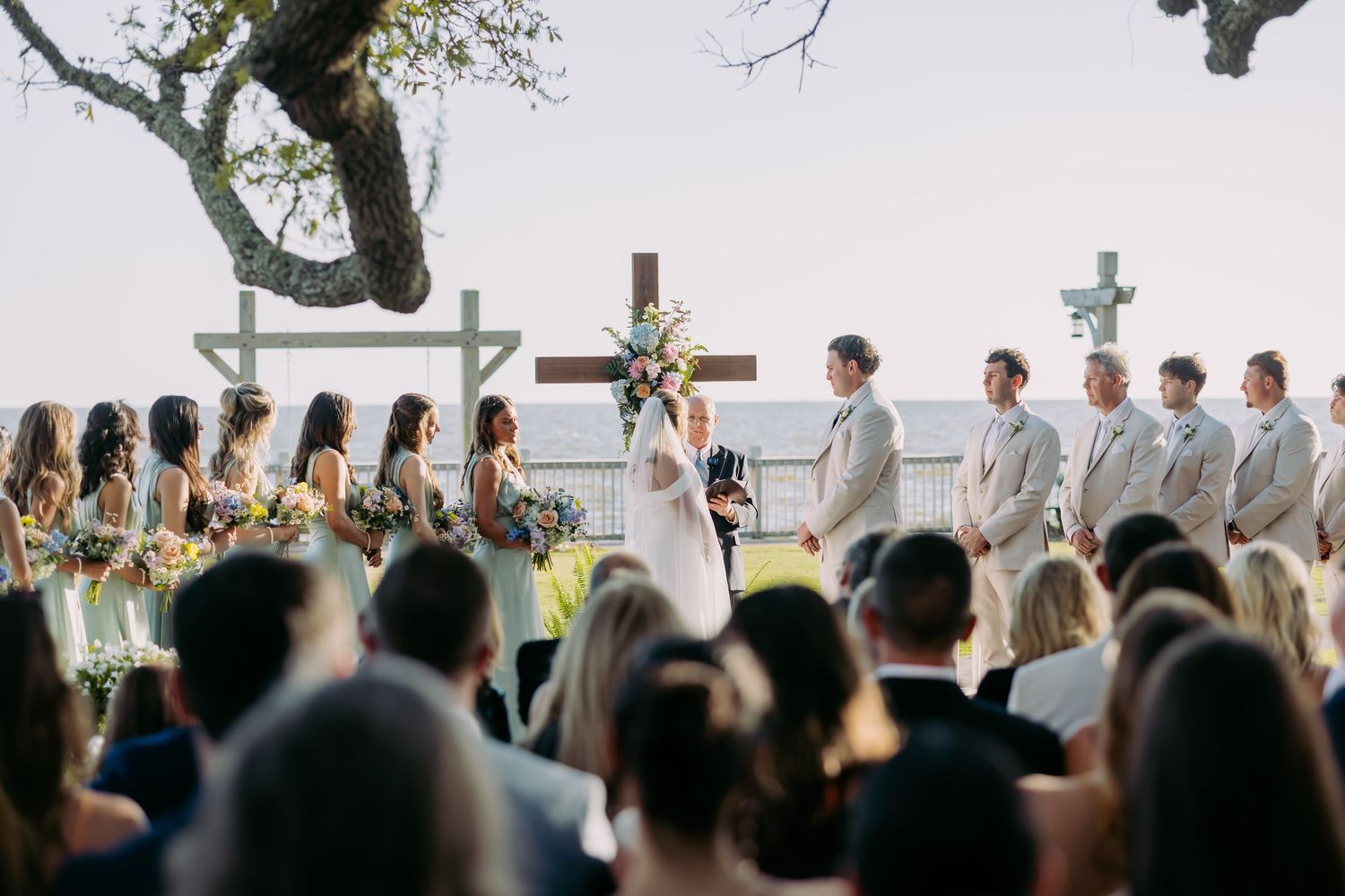 Outdoor wedding ceremony with large cross, bridal party in mint and tan attire by waterfront.